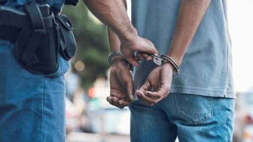 A law enforcement officer in a blue uniform places handcuffs on the wrists of a person wearing a grey shirt and blue jeans.