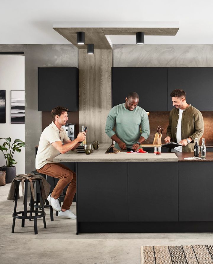 Three men in a modern kitchen, one seated at a counter, two cooking, interacting, and laughing.