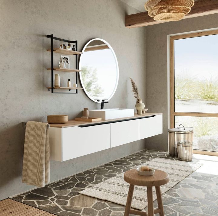 Rustic bathroom with floating vanity, round mirror, wooden stool, and a window overlooking nature.