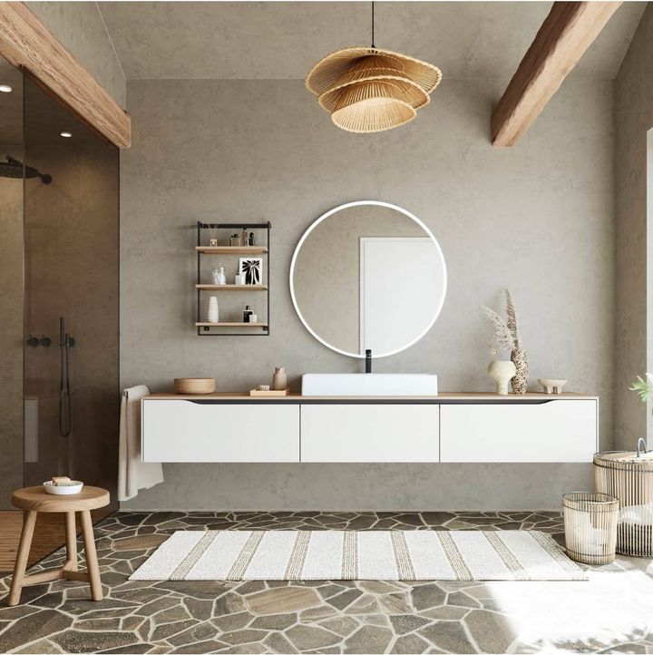 A modern bathroom with a floating vanity, round mirror, and natural stone floor. Neutral tones with wood accents.