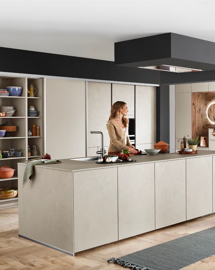 Woman in modern kitchen preparing food at large island. Beige cabinets, black ceiling accent, and wood floors.