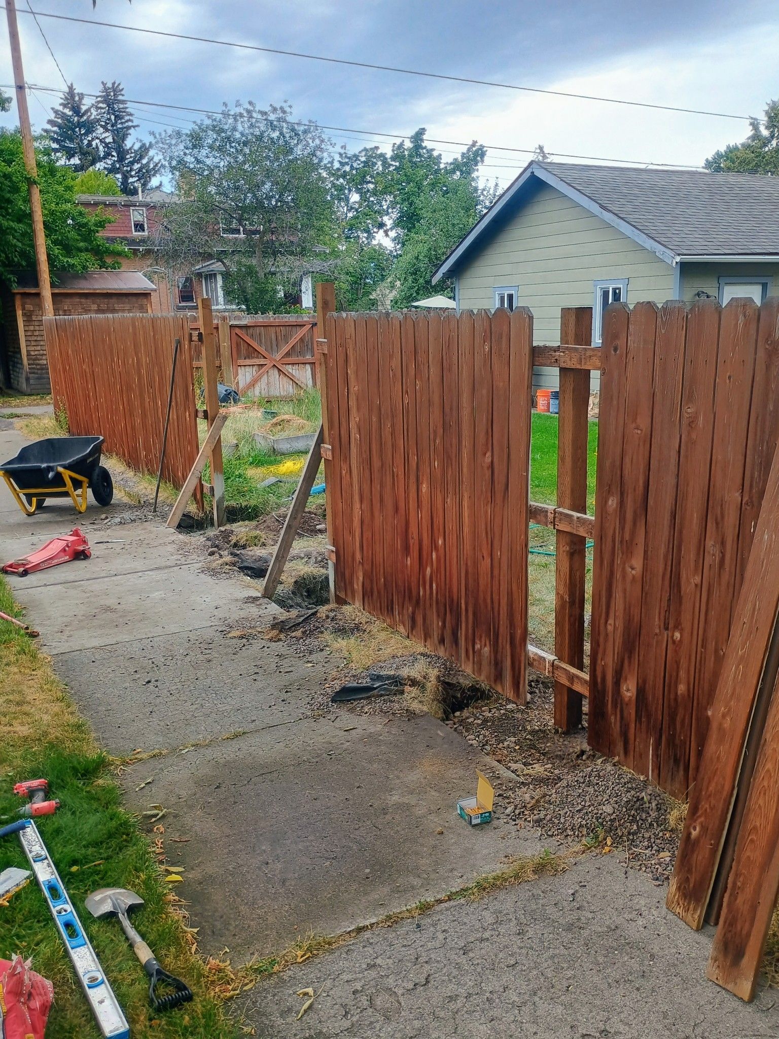 A fence being repaired along a sidewalk. Tools, boards, and debris are scattered around.