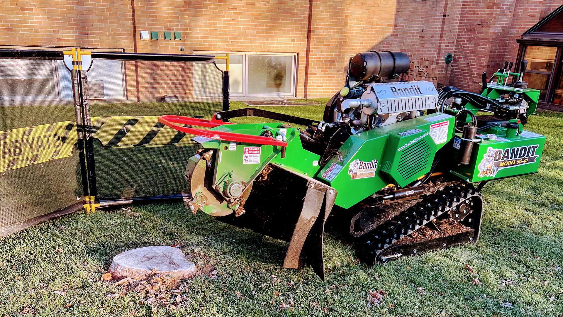 A green stump grinder is sitting in the grass in front of a brick building.