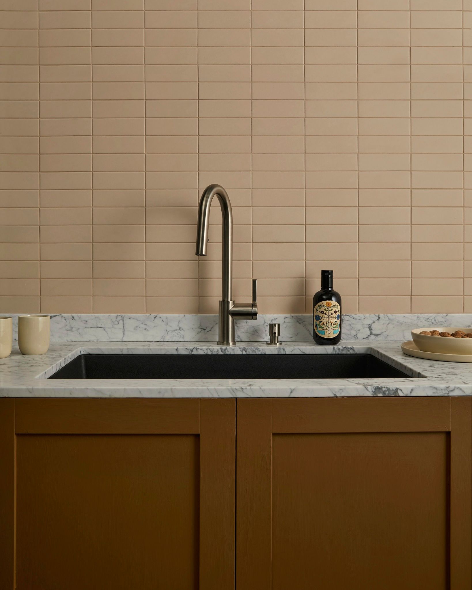 Évier de cuisine avec armoires brunes, comptoir en marbre et dosseret en carrelage beige; bouteille d'huile d'olive sur le comptoir.