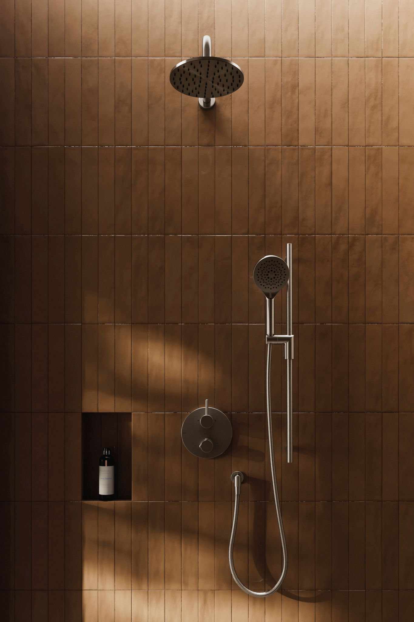 Douche carrelée marron avec pomme de douche, douchette à main et étagère encastrée contenant une bouteille.