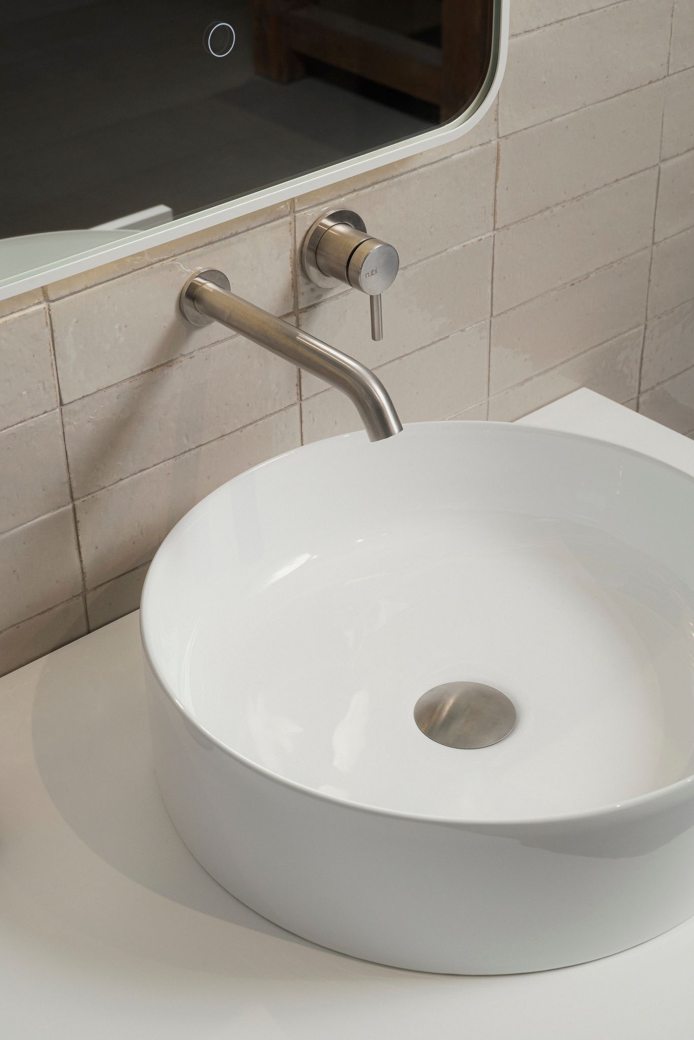 Lavabo de salle de bain blanc et rond avec robinet et bonde chromés, contre un mur carrelé de couleur claire.