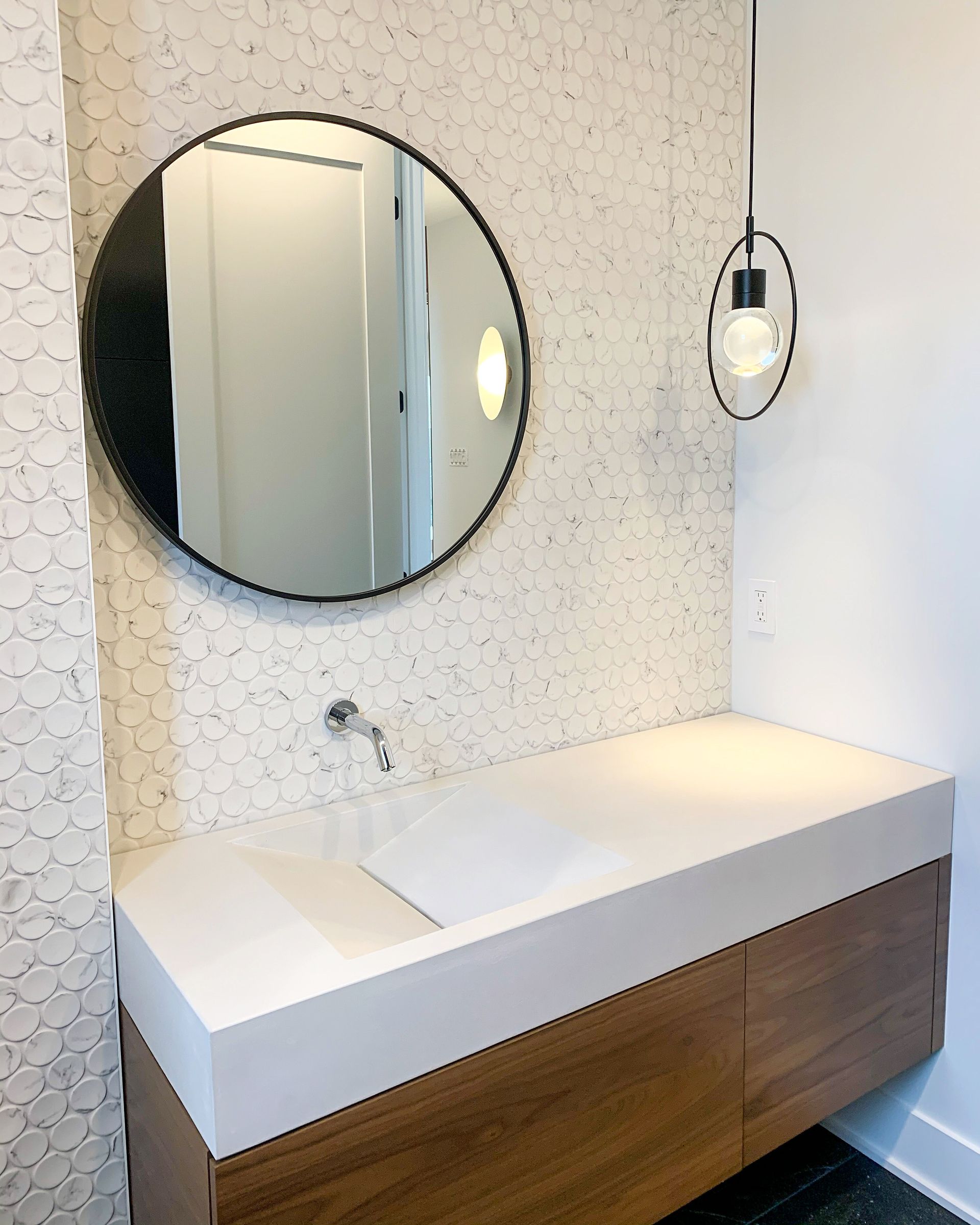 Salle de bain moderne avec vanité flottante en bois, comptoir blanc, miroir rond noir et dosseret en carrelage hexagonal.
