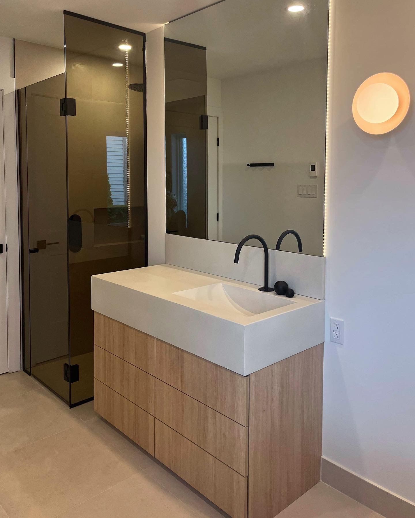 Salle de bain moderne avec vanité en bois clair, grand miroir et cabine de douche en verre.