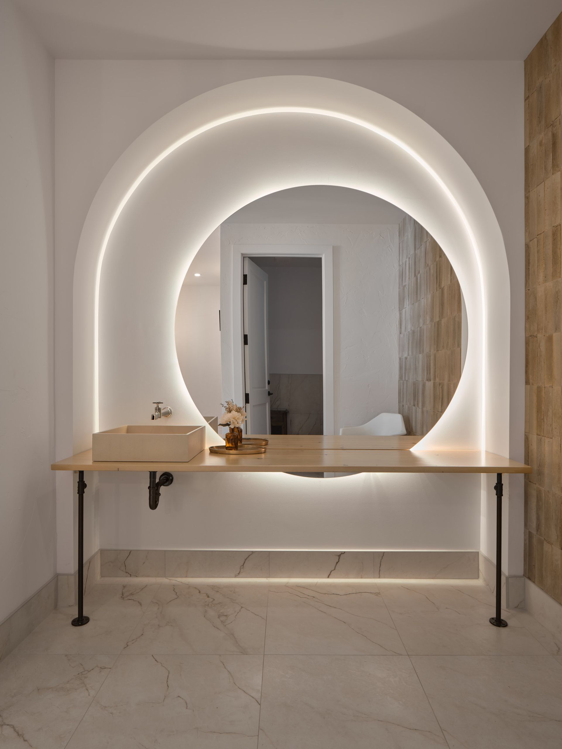 Salle de bain moderne avec miroir rond, comptoir et lavabo en bois, éclairage en arc et murs carrelés.