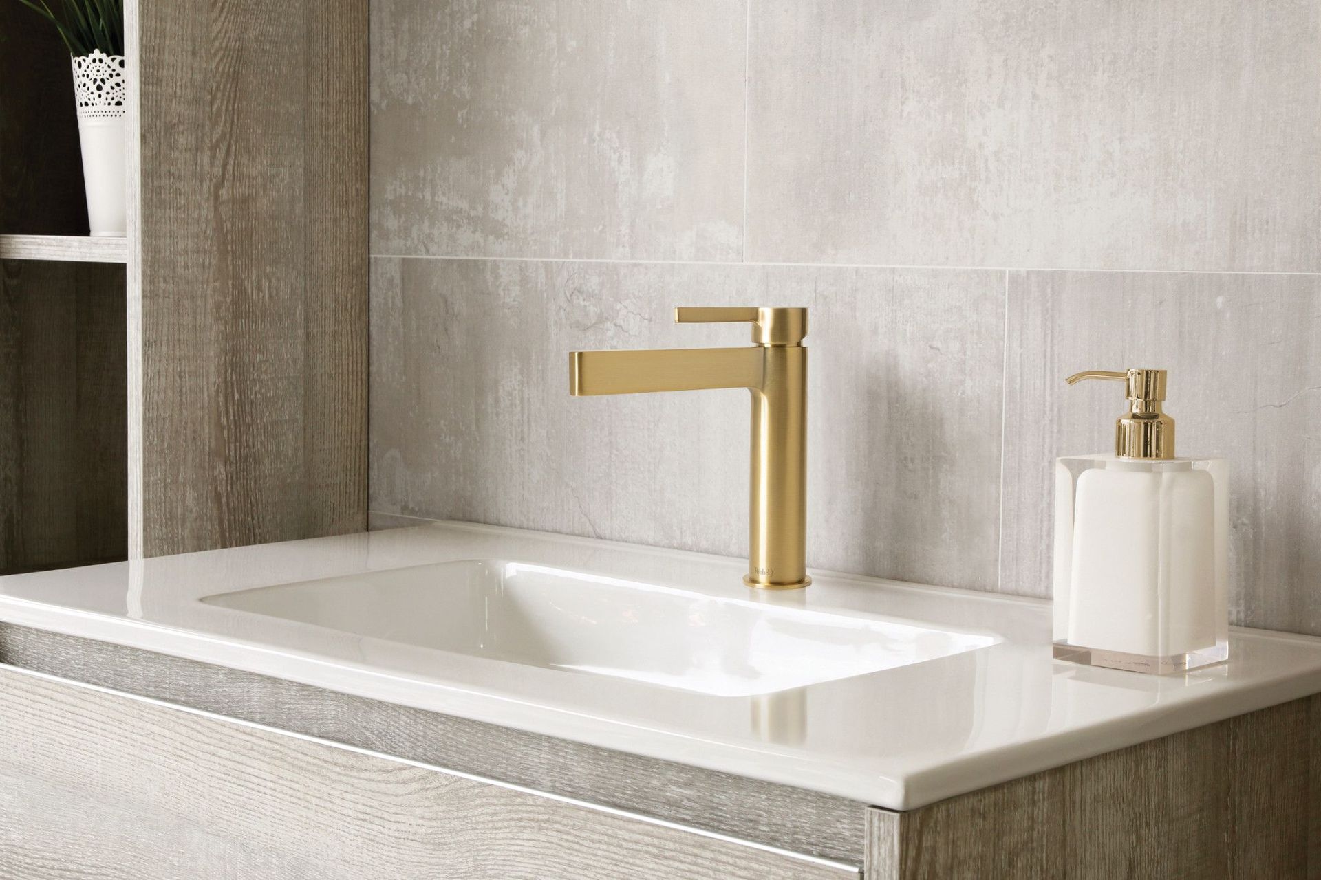 Robinet doré sur un lavabo blanc. Carrelage en pierre grise, meubles en bois et distributeur de savon.