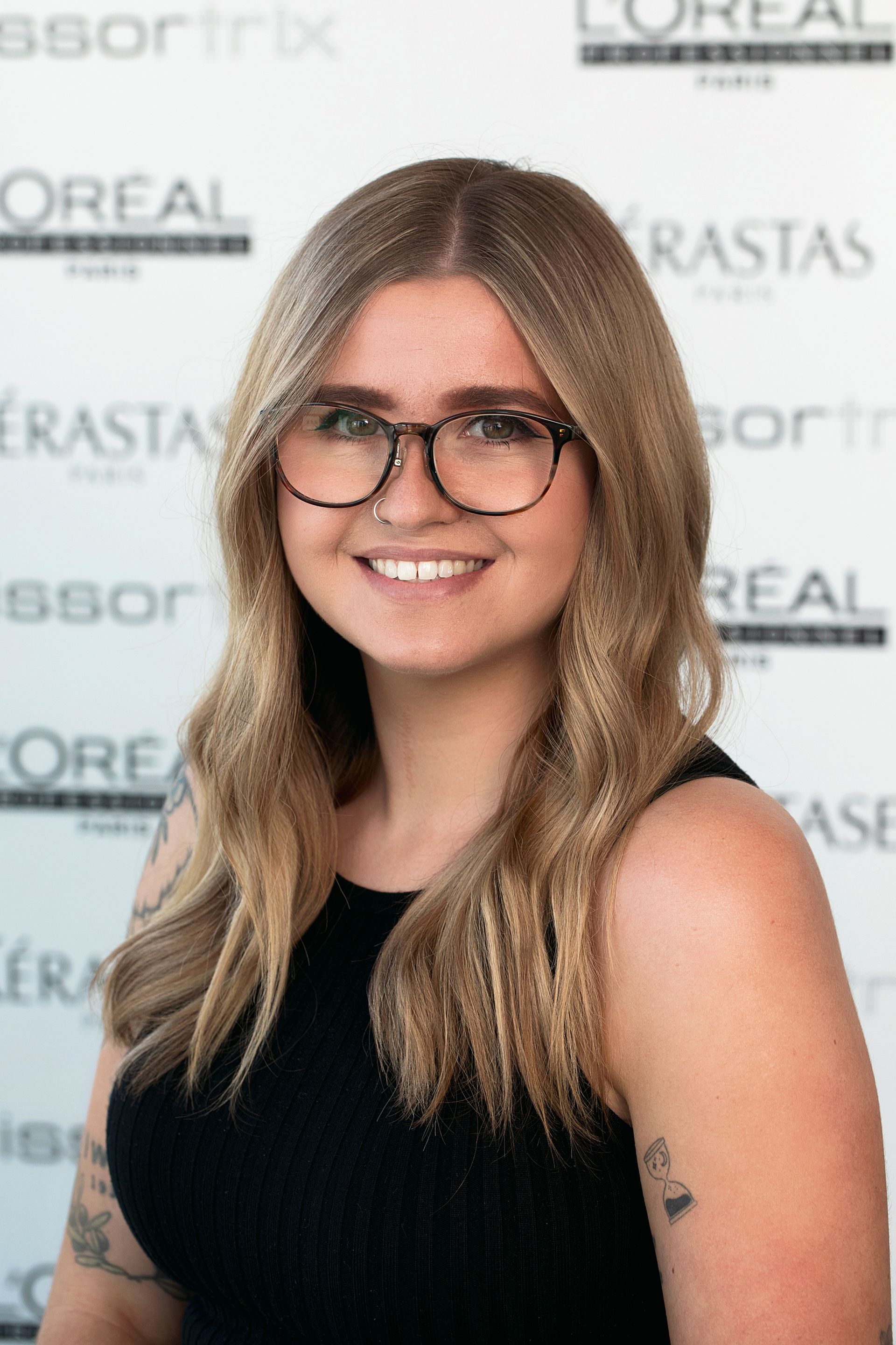 A woman wearing glasses and a black shirt is smiling in front of a sign that says l' oreal