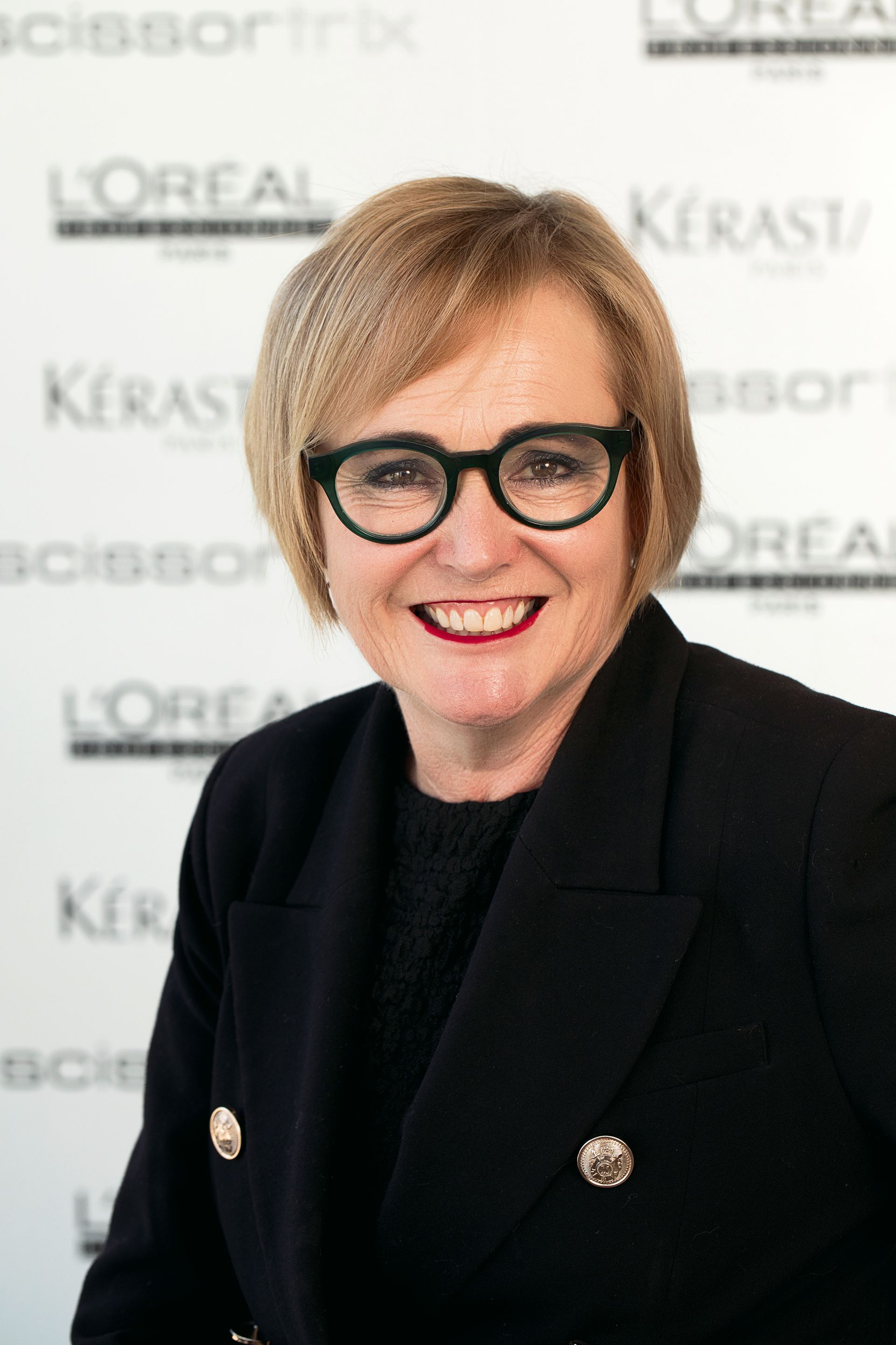 A woman wearing glasses is smiling in front of a kerastase sign