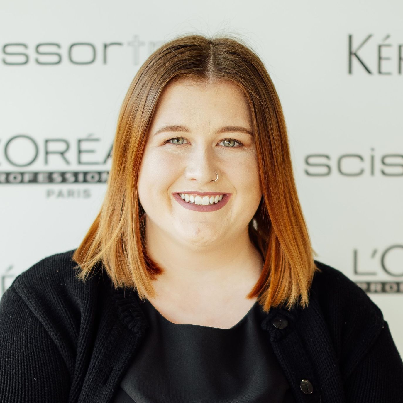 A woman wearing glasses and a black shirt is smiling in front of a sign that says l' oreal