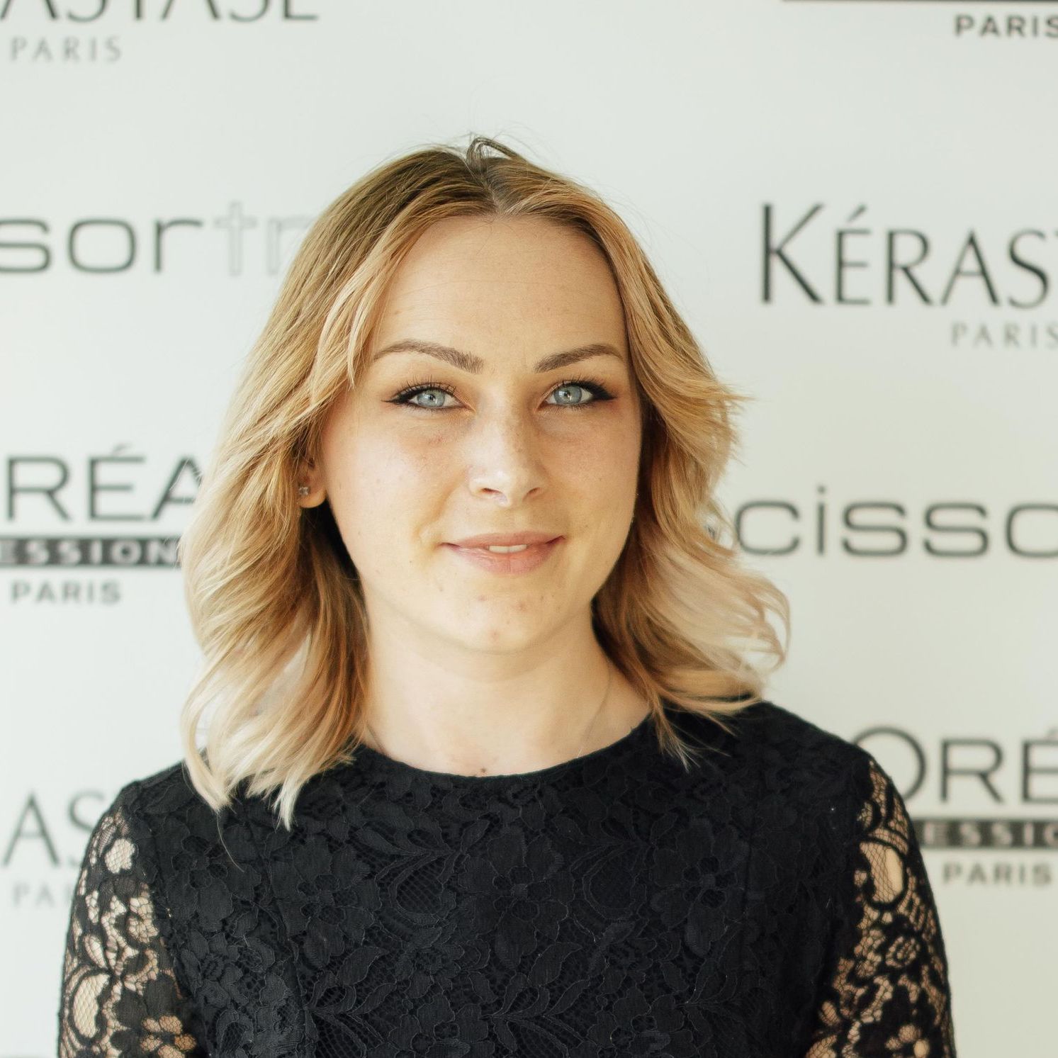 A woman wearing glasses and a black shirt is smiling in front of a sign that says l' oreal