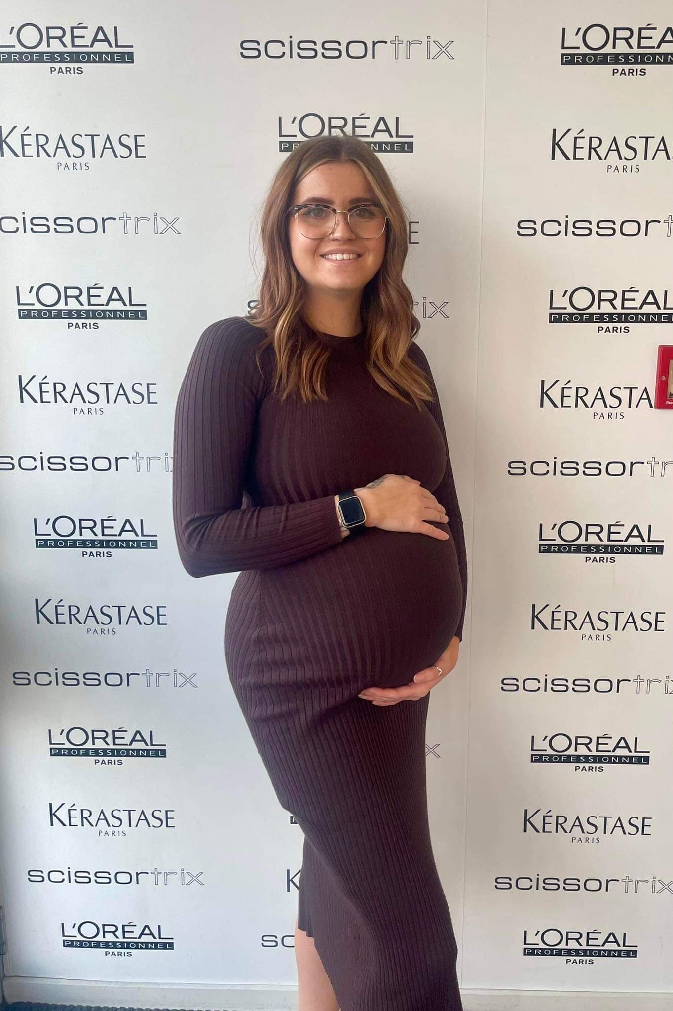 A pregnant woman is standing in front of a wall holding her belly.