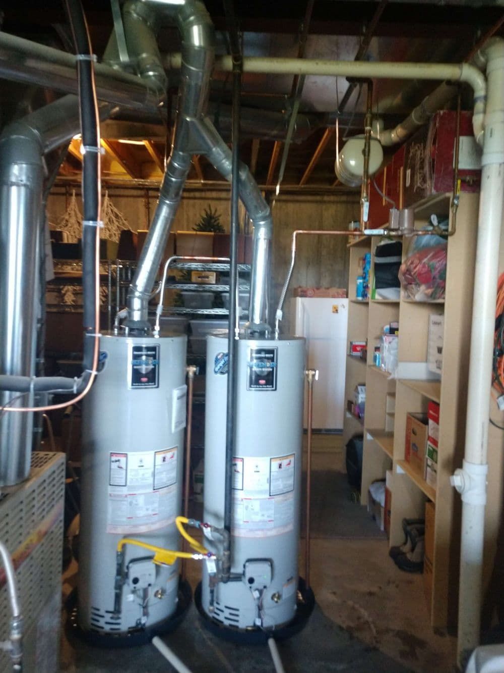Two water heaters side-by-side in a basement. They have silver exhaust pipes. Shelves and pipes are visible.