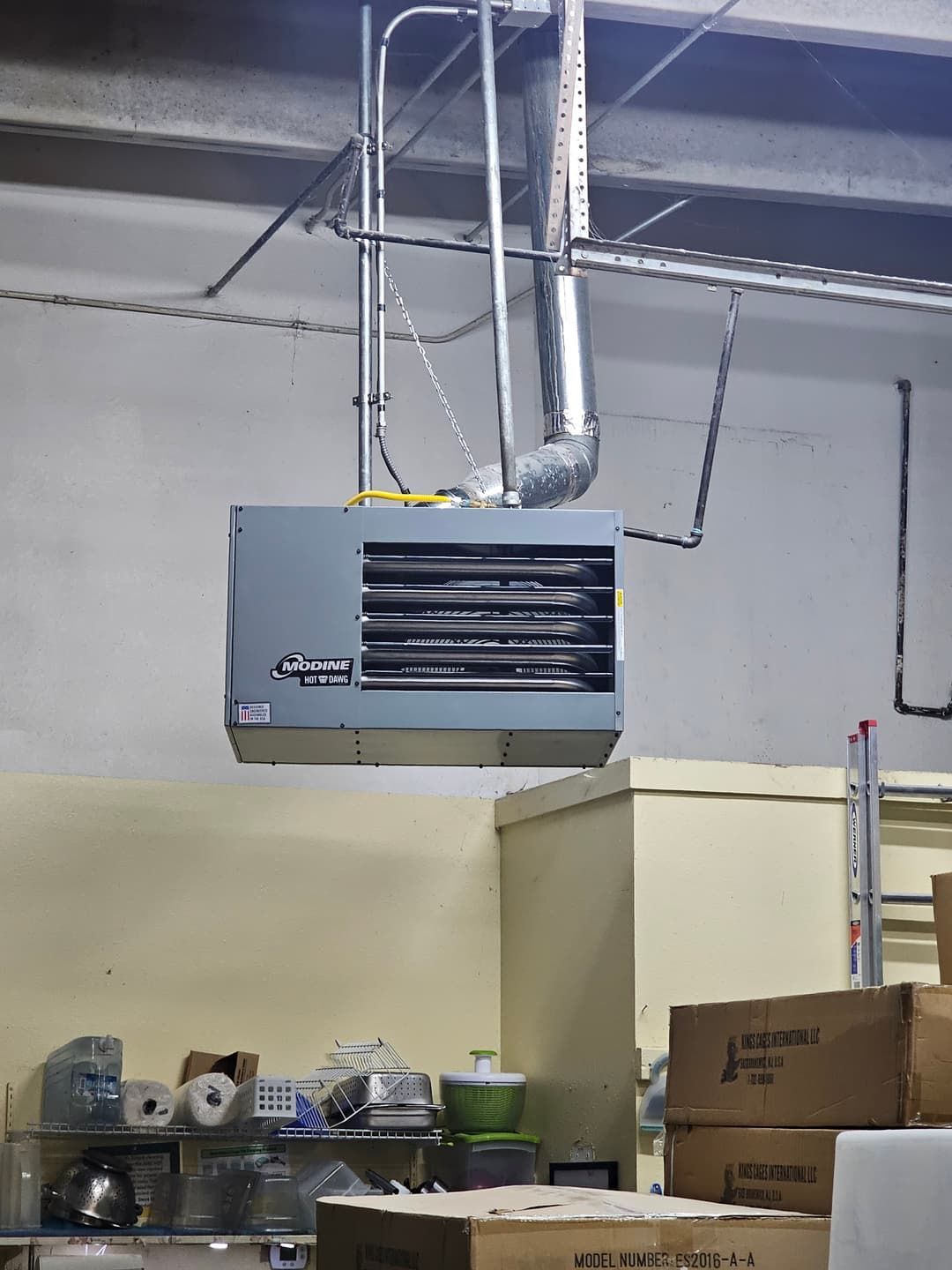 Gray overhead gas heater in a warehouse setting with ductwork attached.