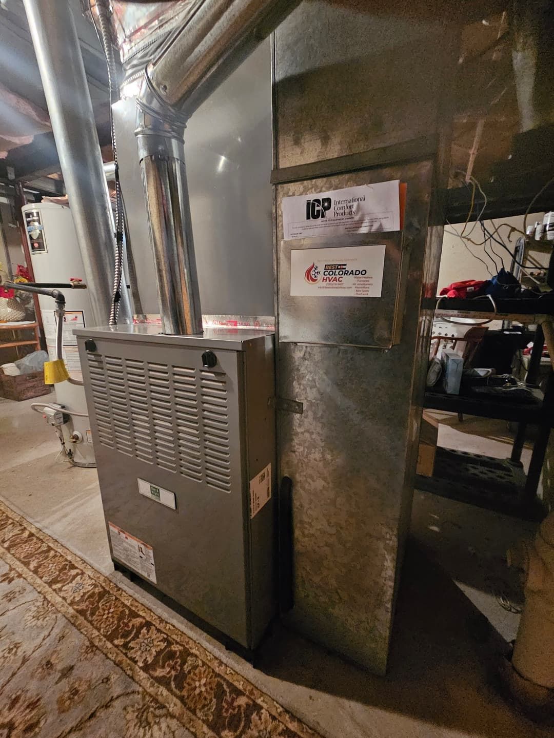Furnace unit in a basement setting, with silver vents, a rug, and service stickers.