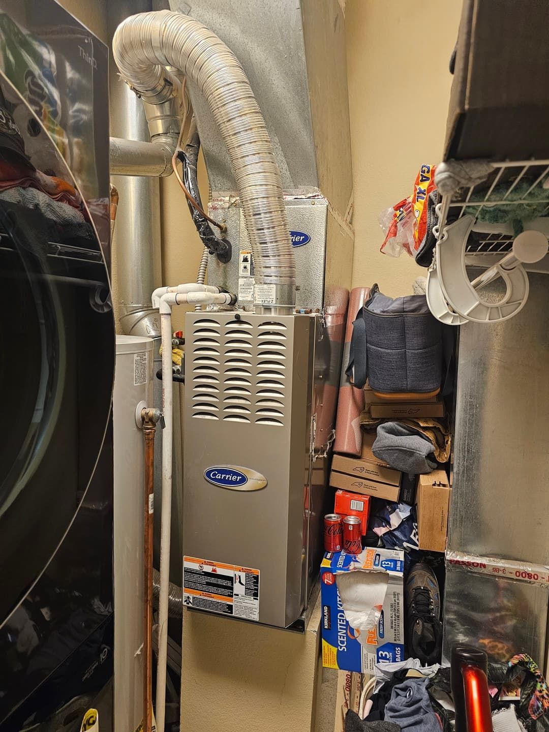 Furnace in a utility room with flexible ductwork. Shelf cluttered with cleaning supplies next to it.