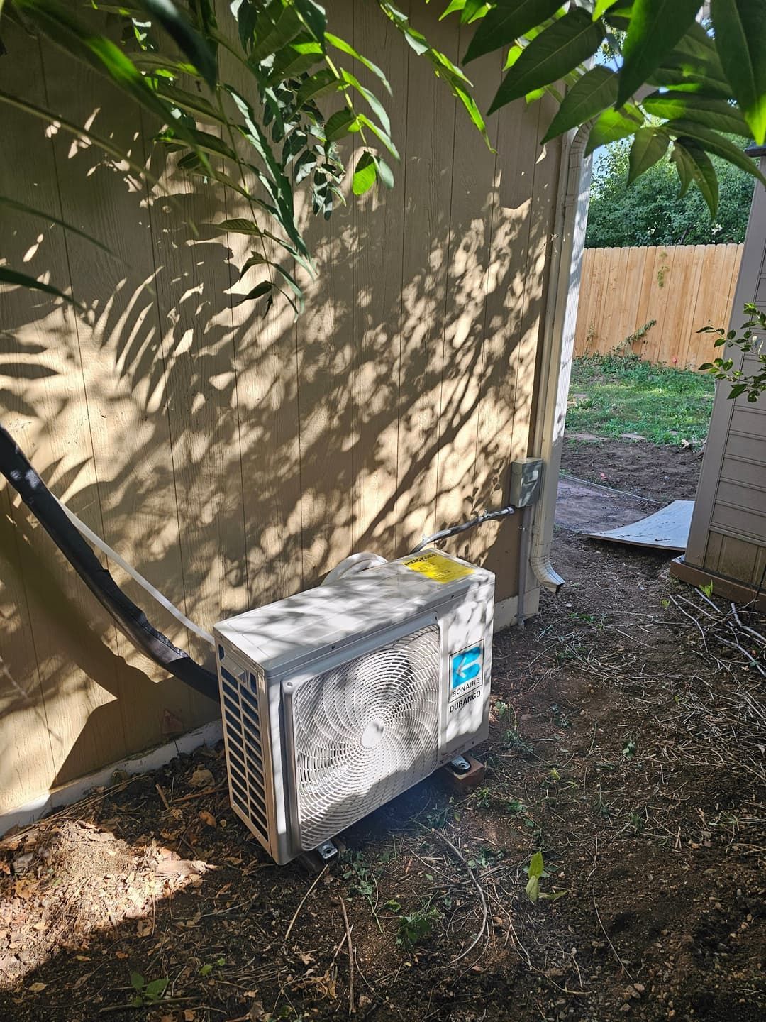 Outdoor AC unit against a wall, shadowed by leaves, with a grassy area and fence visible.