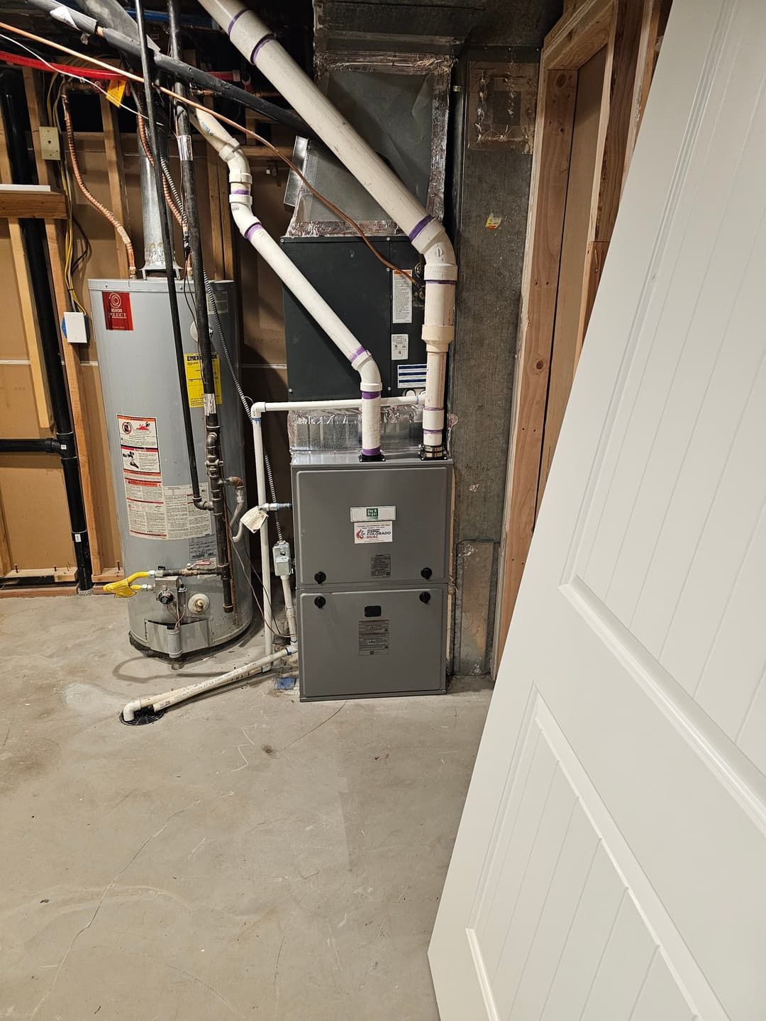 A basement with a gray furnace, a white water heater, and white PVC pipes.