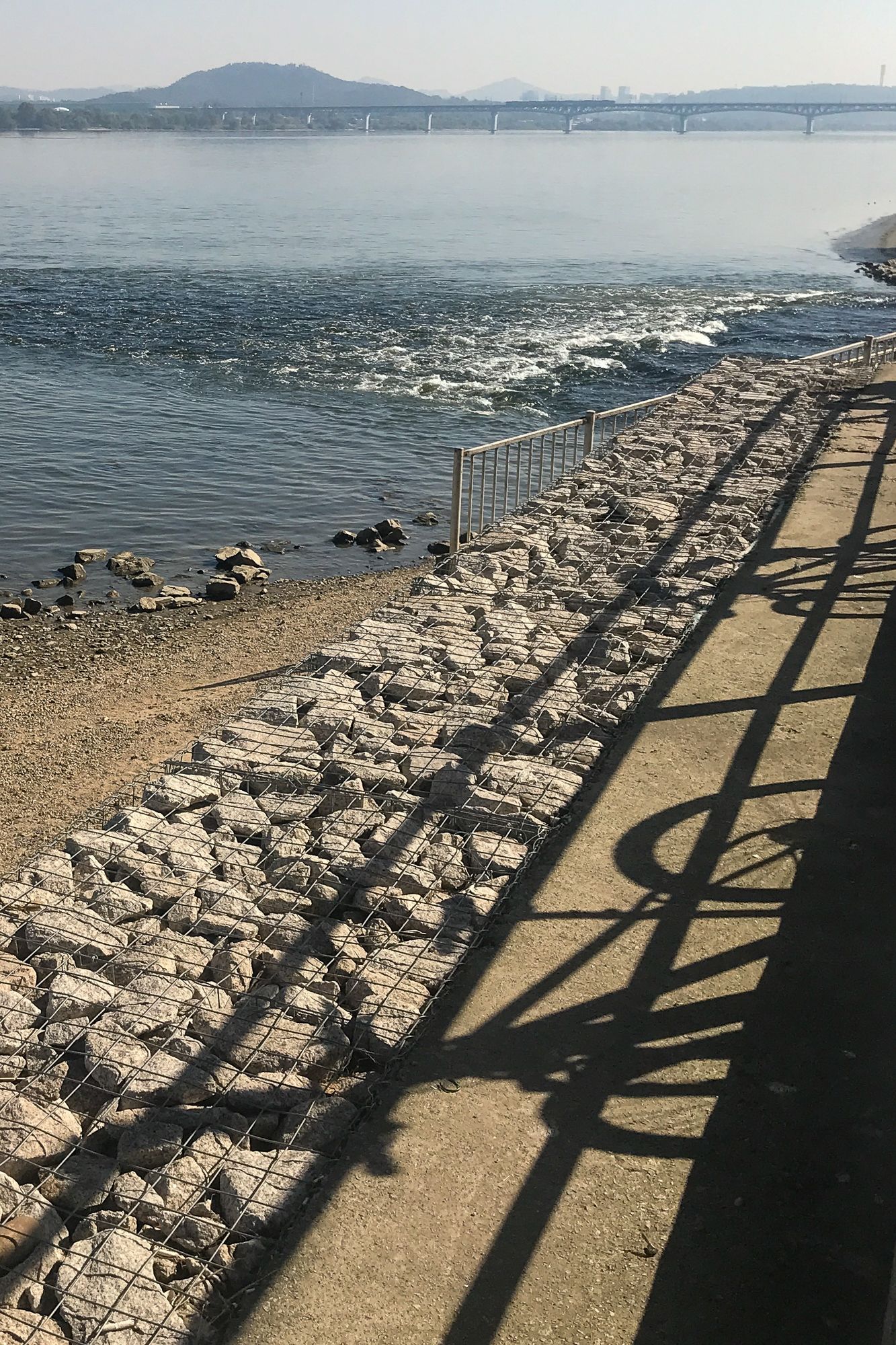 A shadow of a person walking on a sidewalk next to a body of water.