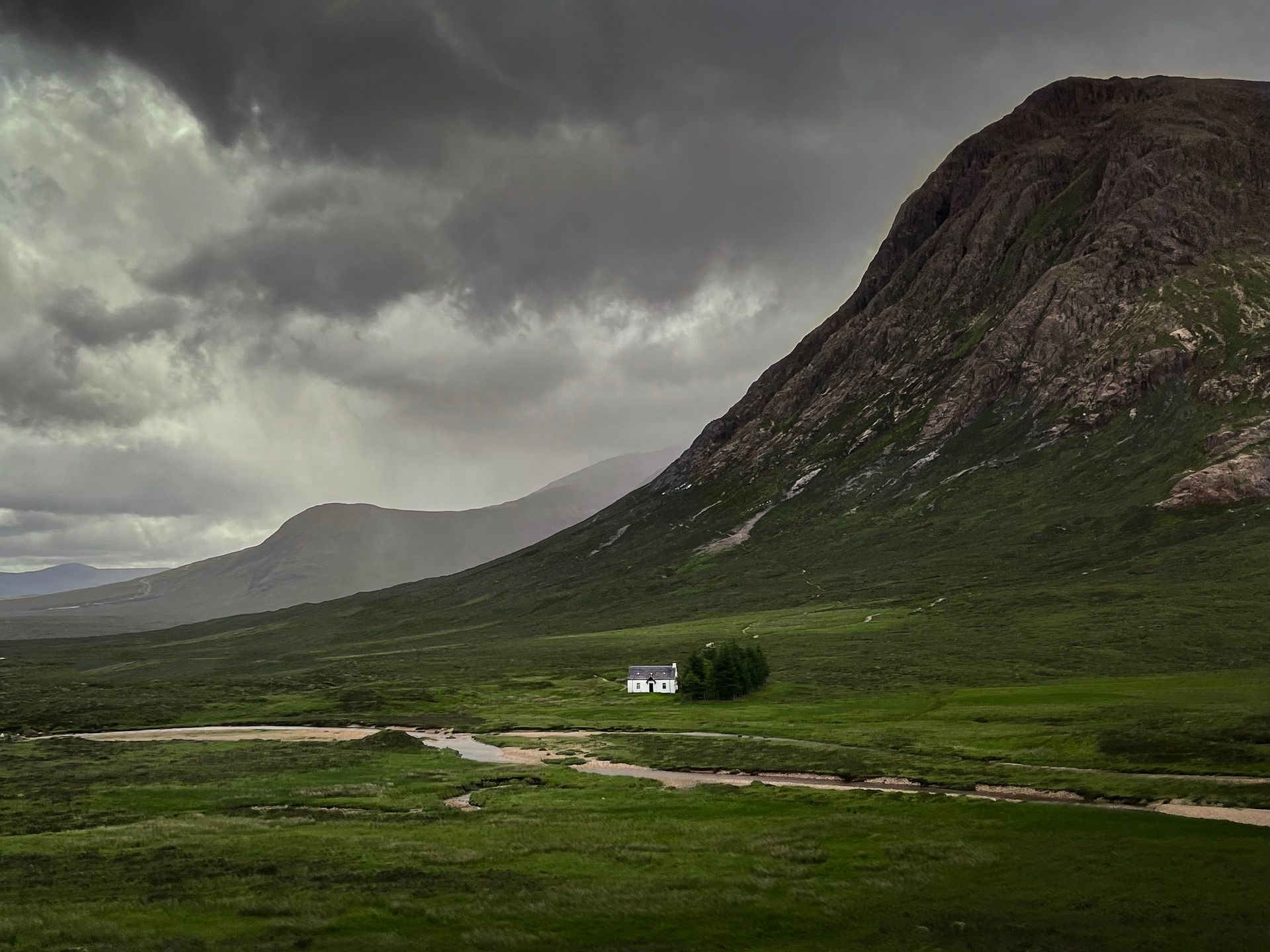 A small house is sitting in the middle of a grassy field next to a mountain.