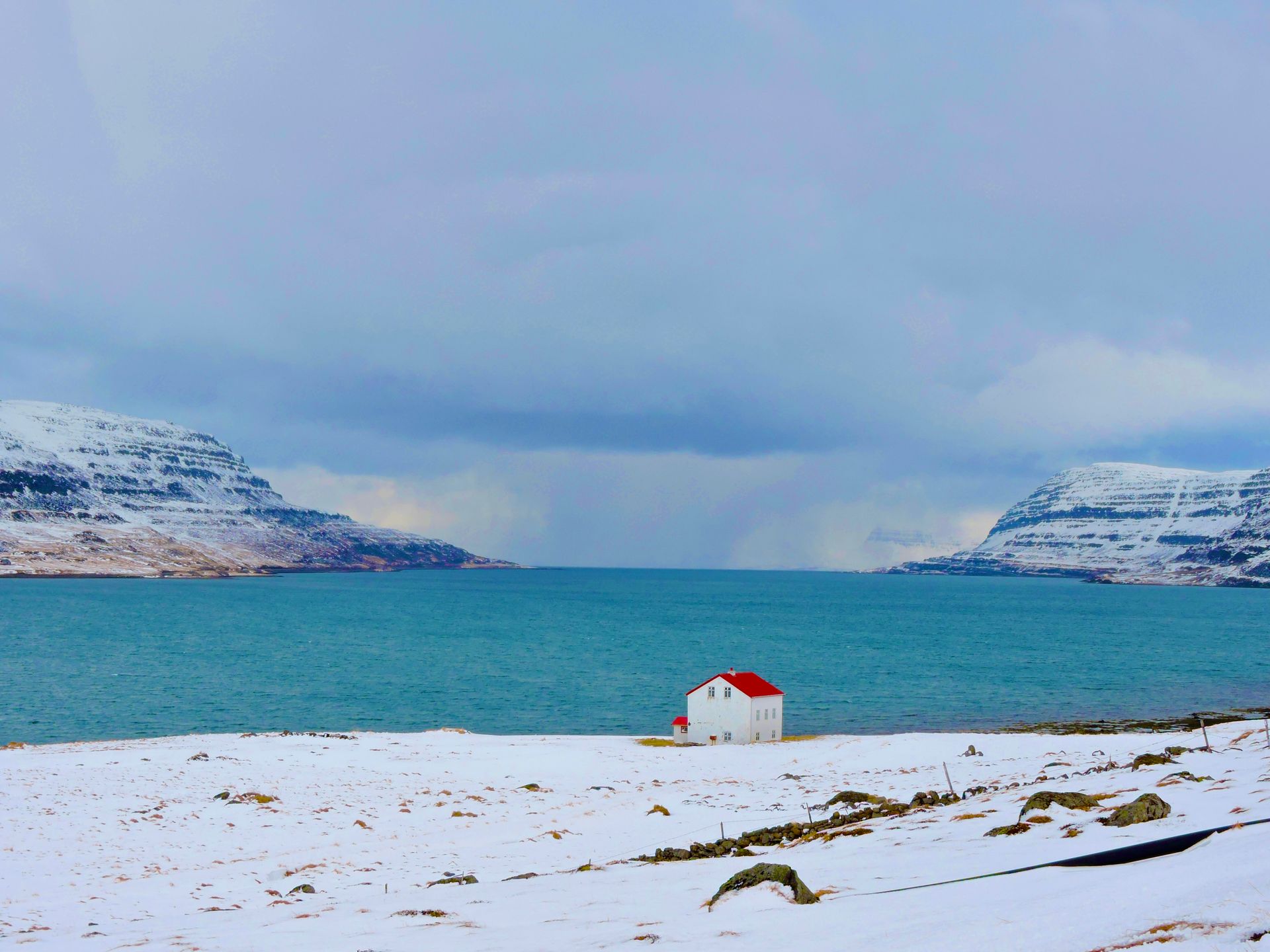 A small white house with a red roof sits on a snowy beach near the ocean.