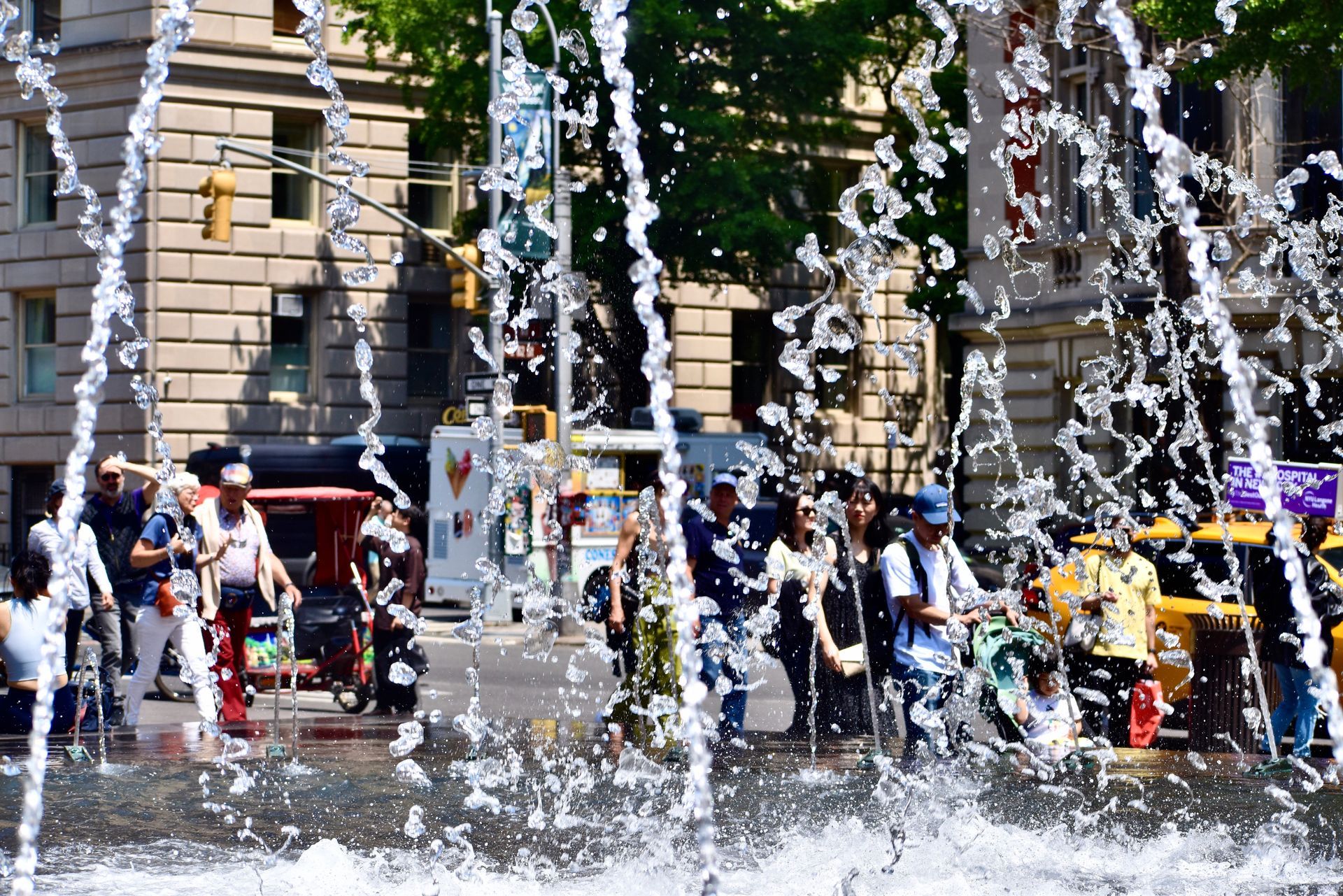 A group of people are standing in front of a fountain in a city.