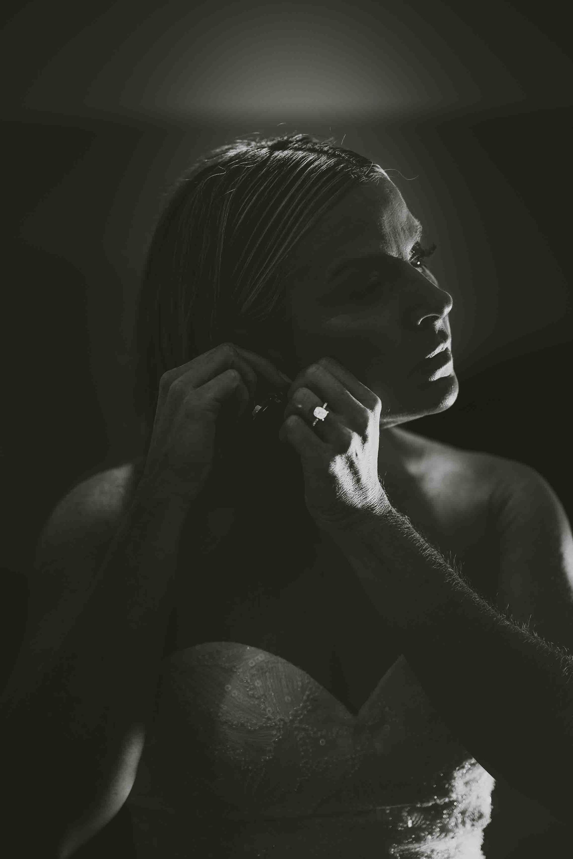 A woman in a wedding dress is putting on her earrings in a black and white photo.