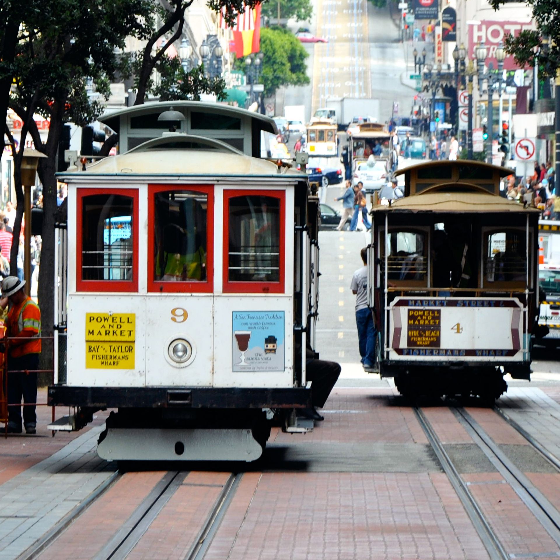 Two trolleys are going down a street with a hot sign in the background