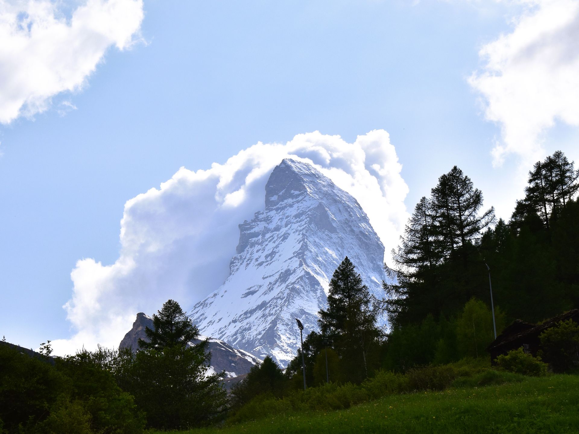 A snowy mountain with trees in the foreground and a blue sky with clouds