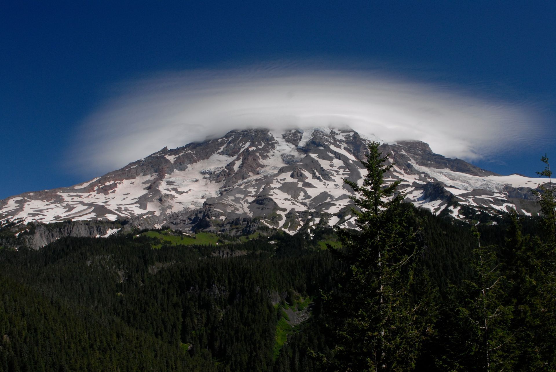 A mountain covered in snow and clouds with trees in the foreground