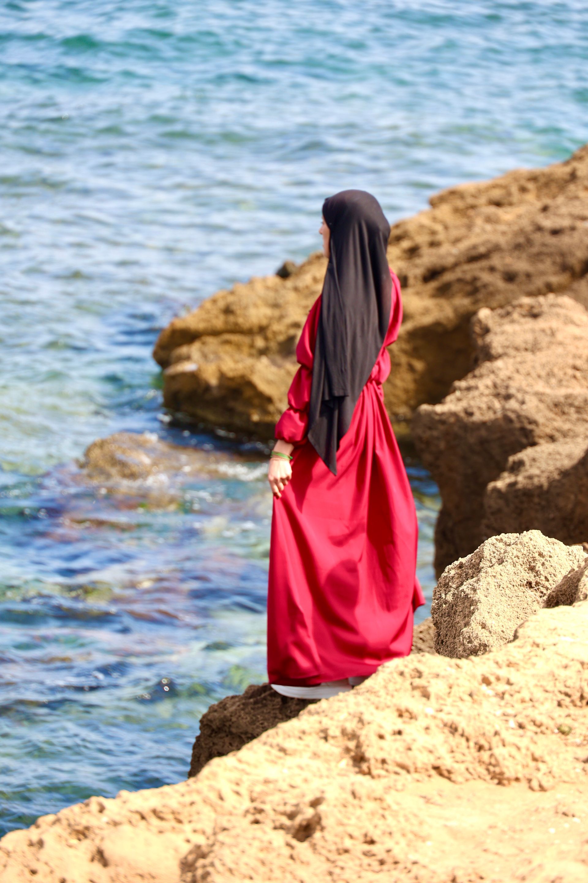 A woman in a red dress is standing on a rock near the ocean.