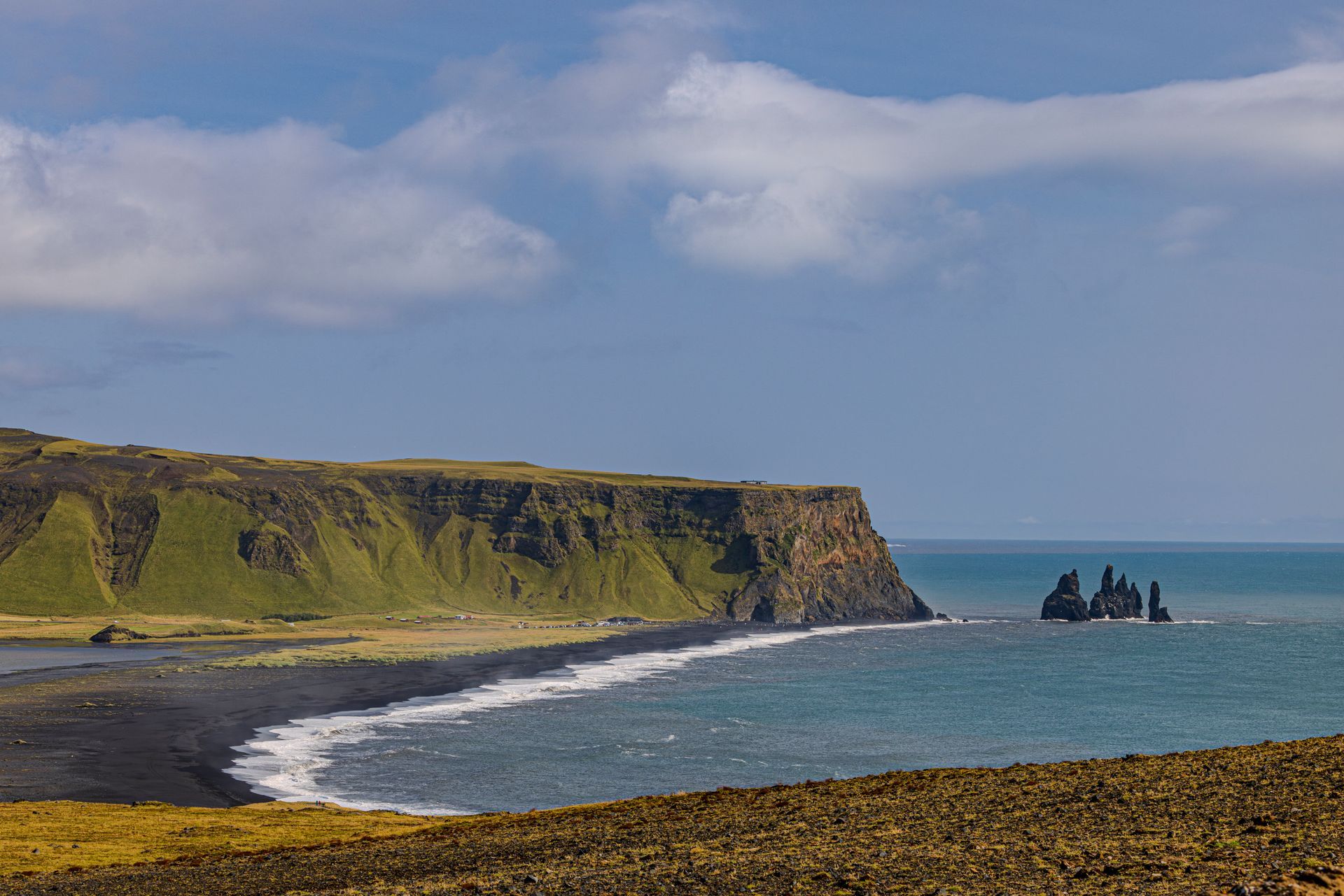A black sand beach with a cliff in the background and a large rock formation in the ocean.