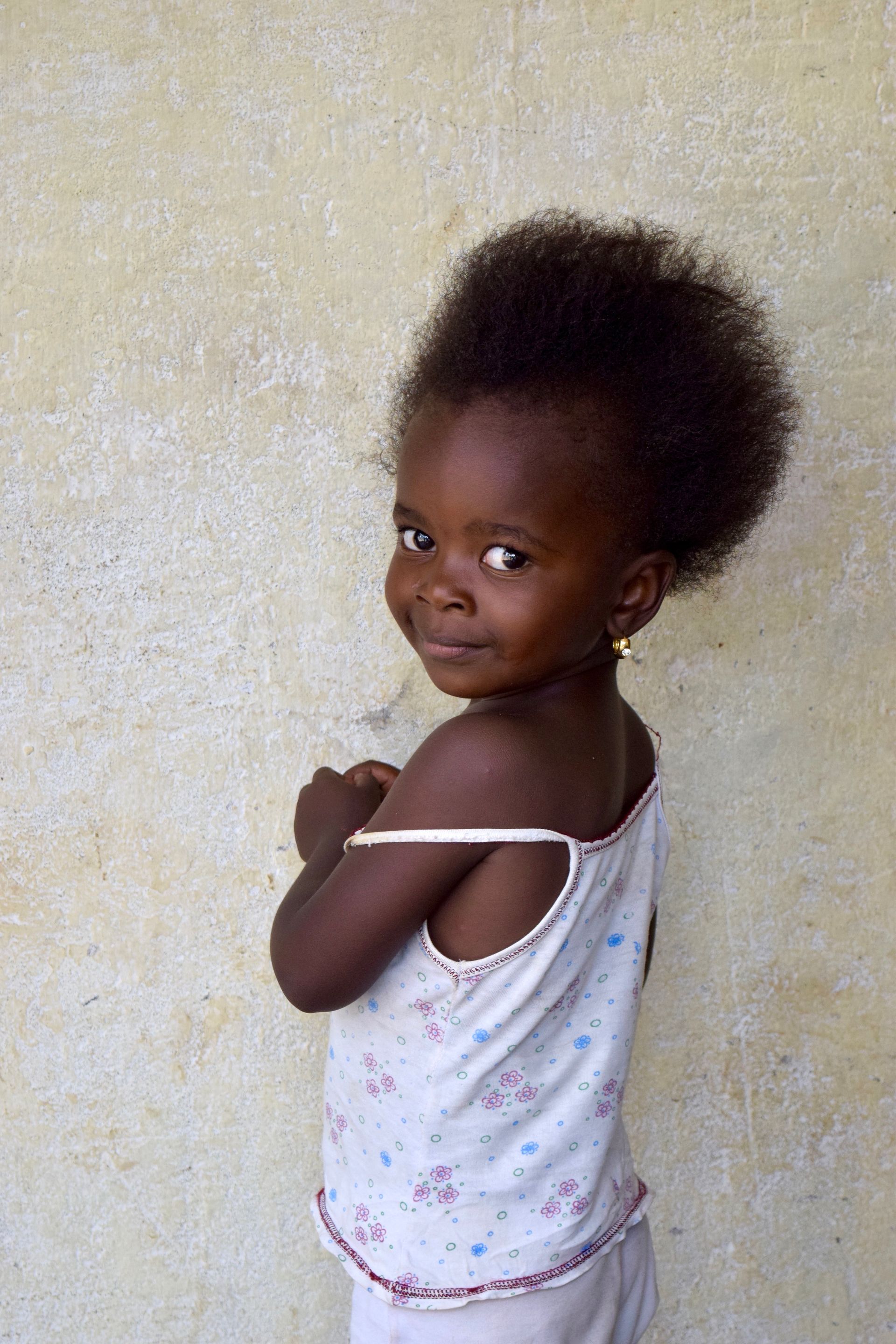 A little girl in a white tank top is standing in front of a wall.