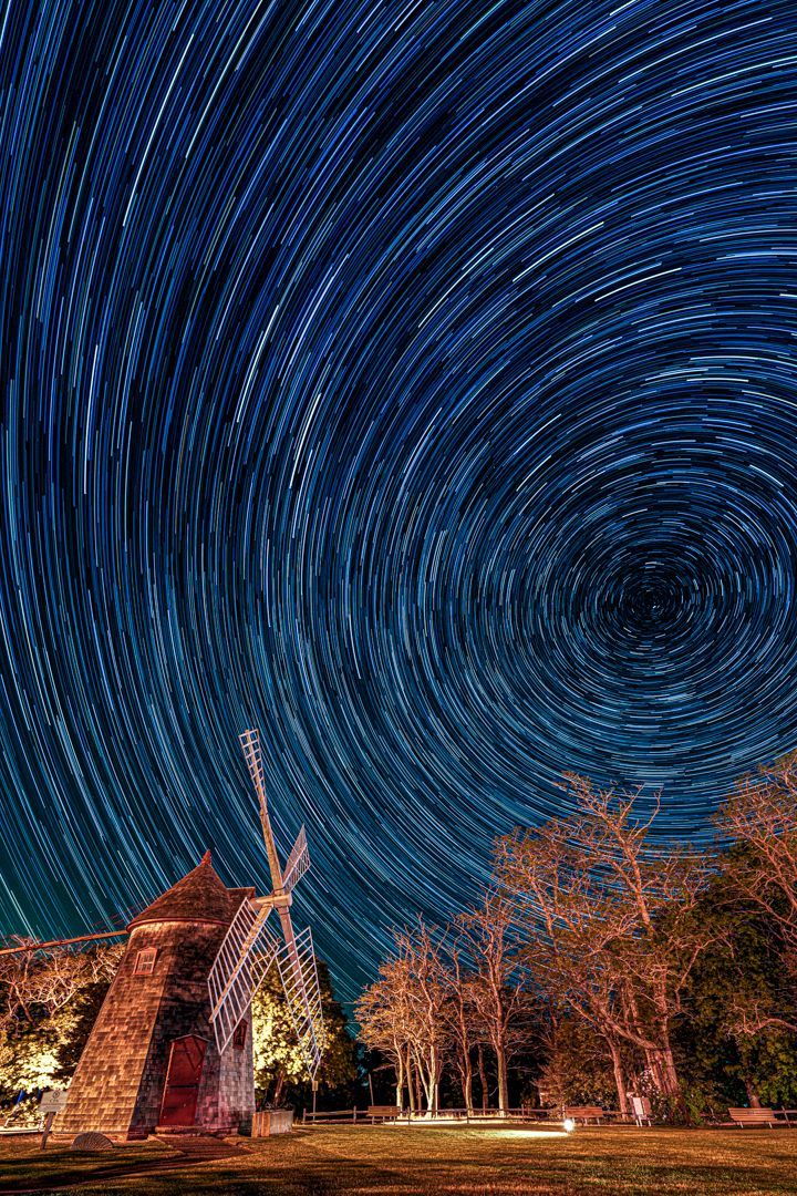 A windmill is surrounded by trees under a starry night sky.