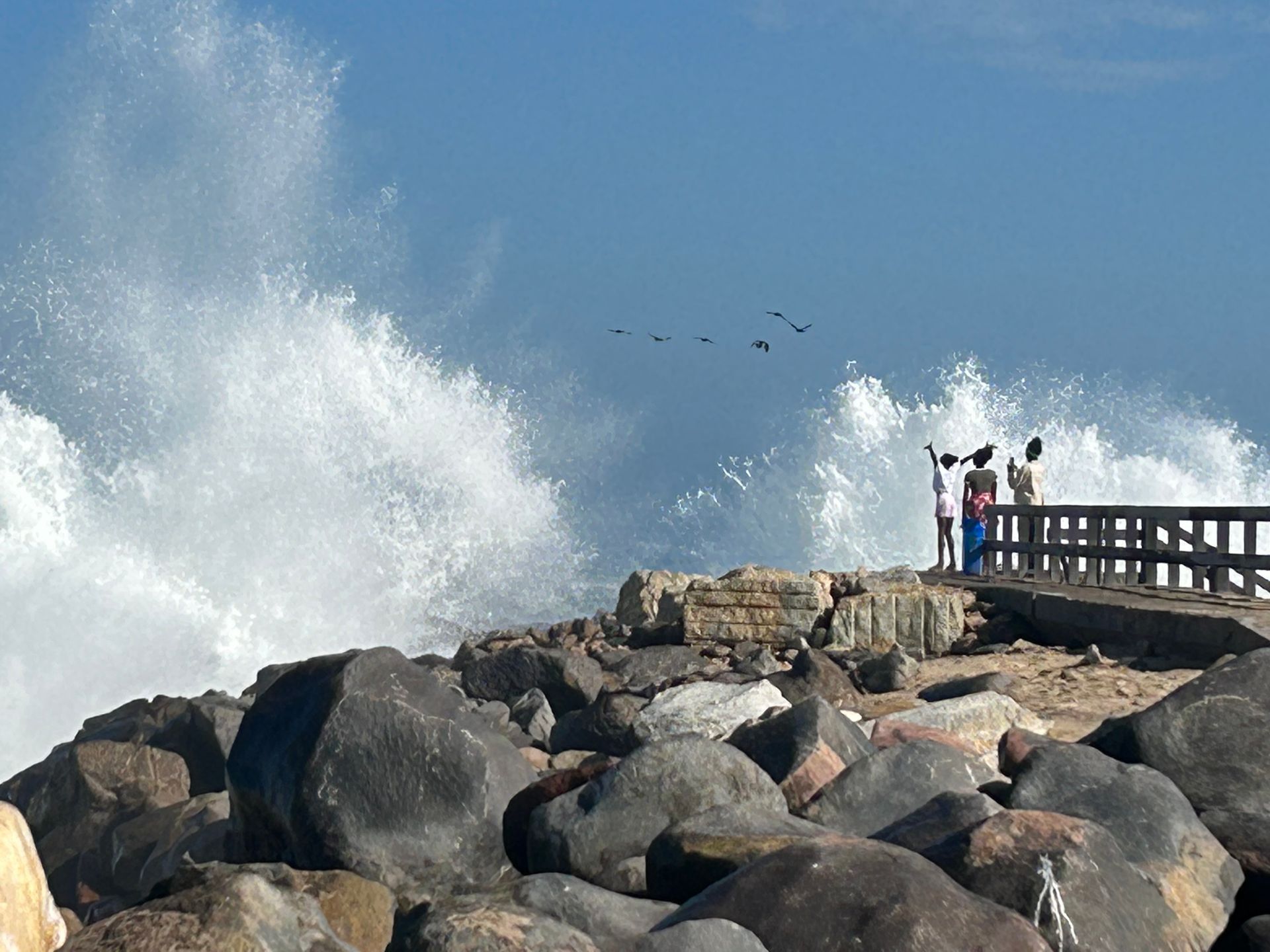 a couple standing on a pier looking at waves crashing on the rocks