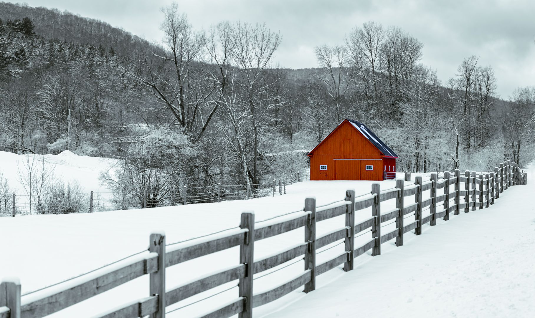 A red barn is sitting in the middle of a snowy field next to a wooden fence.