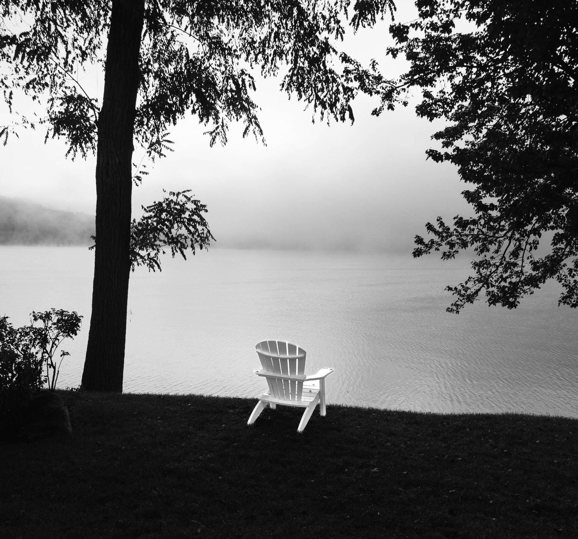 A black and white photo of a lake with a chair in the foreground