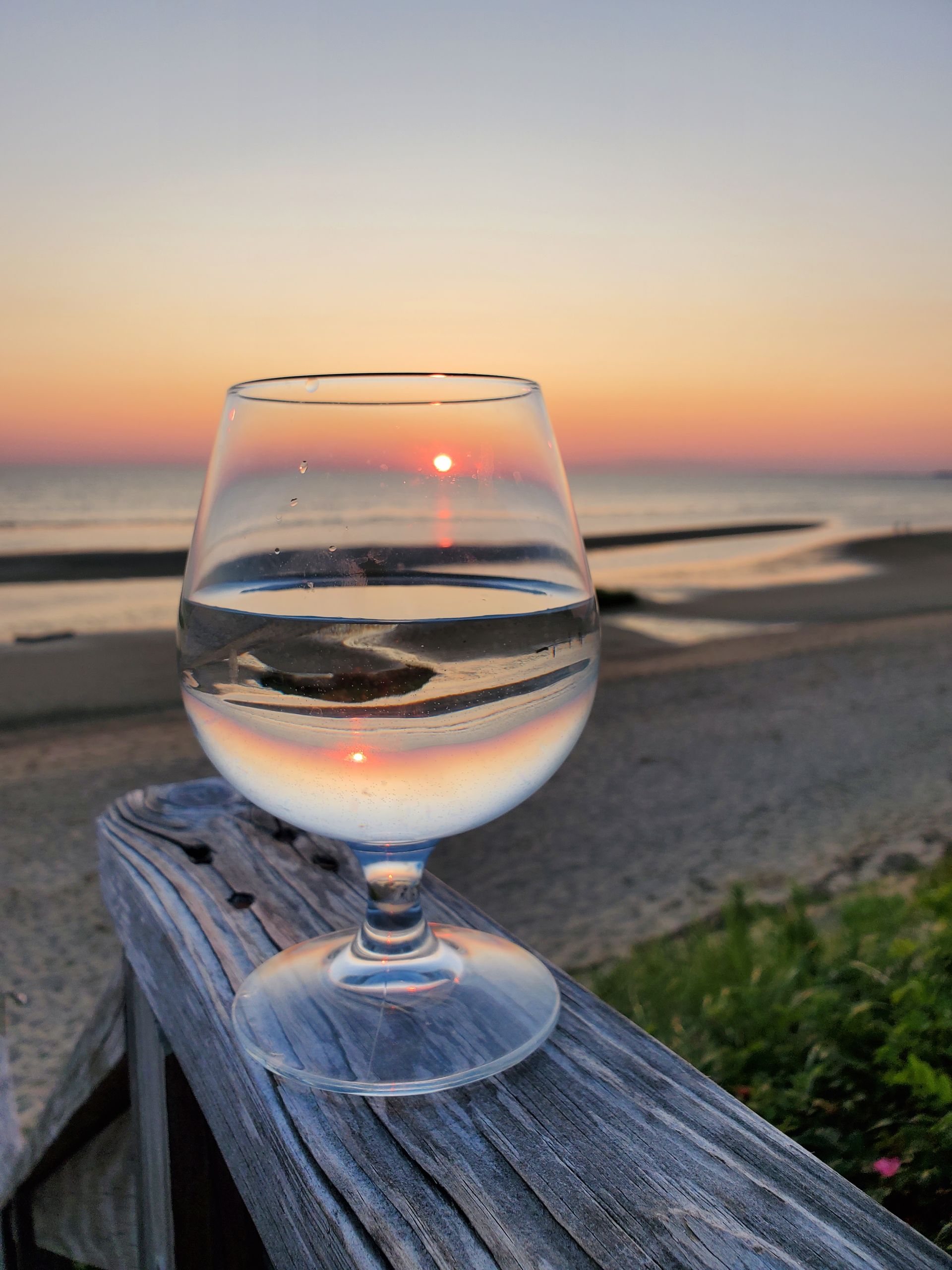 a glass of water sits on a wooden railing overlooking the ocean at sunset