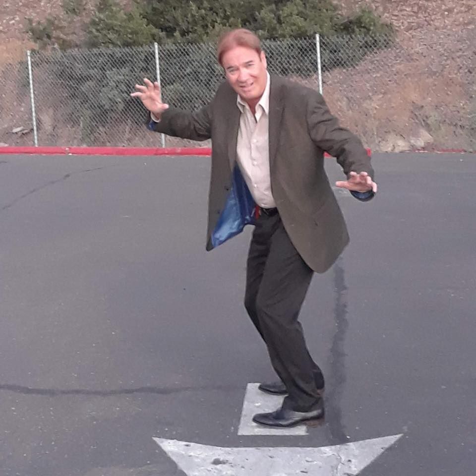Man posing playfully on a painted parking lot arrow, smiling with arms outstretched.