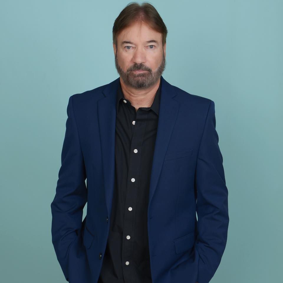 Man in a navy blazer over a black shirt standing against a light blue background