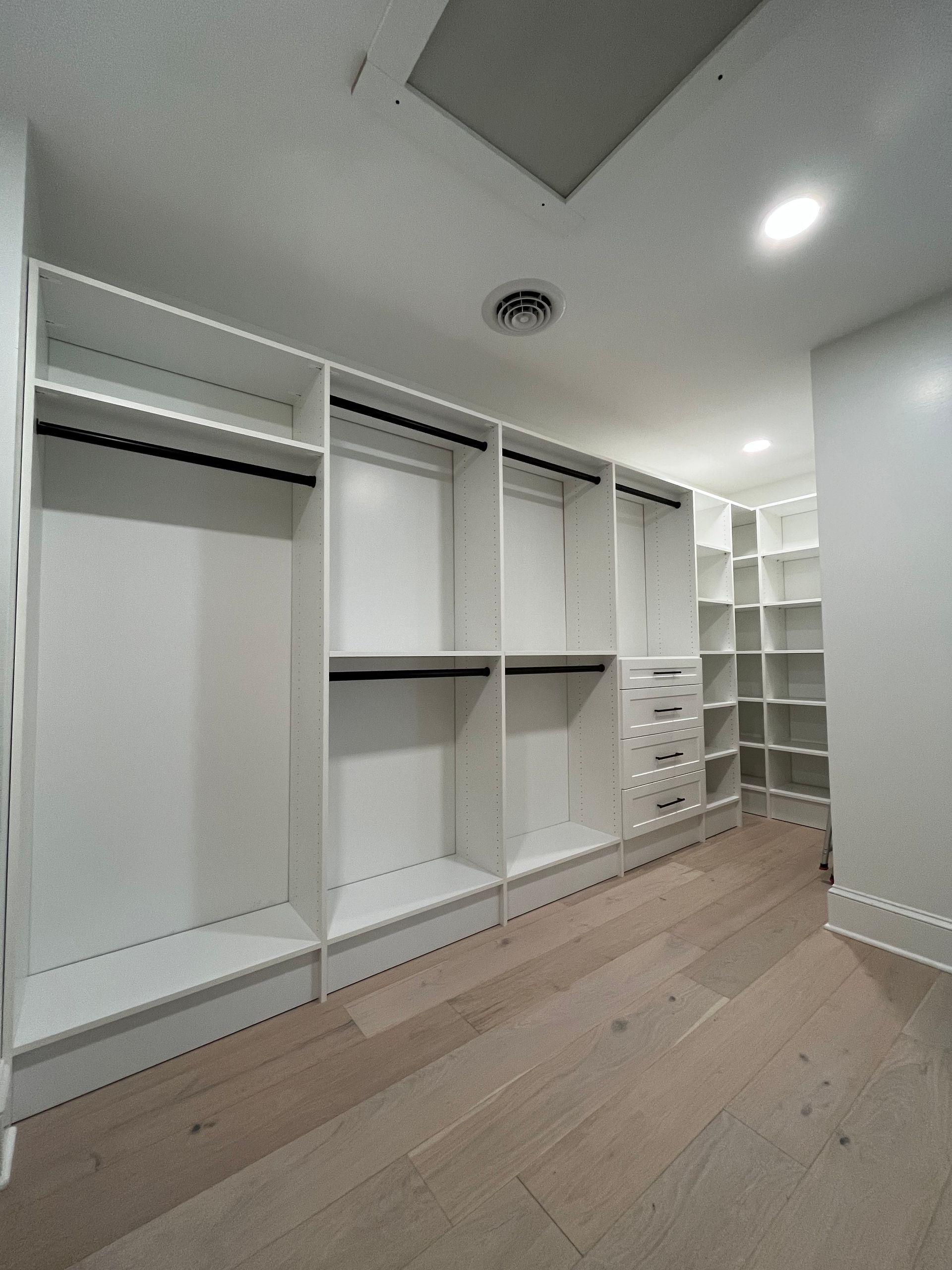 White walk-in closet with built-in shelving, drawers, and hanging rods on light wood floor.