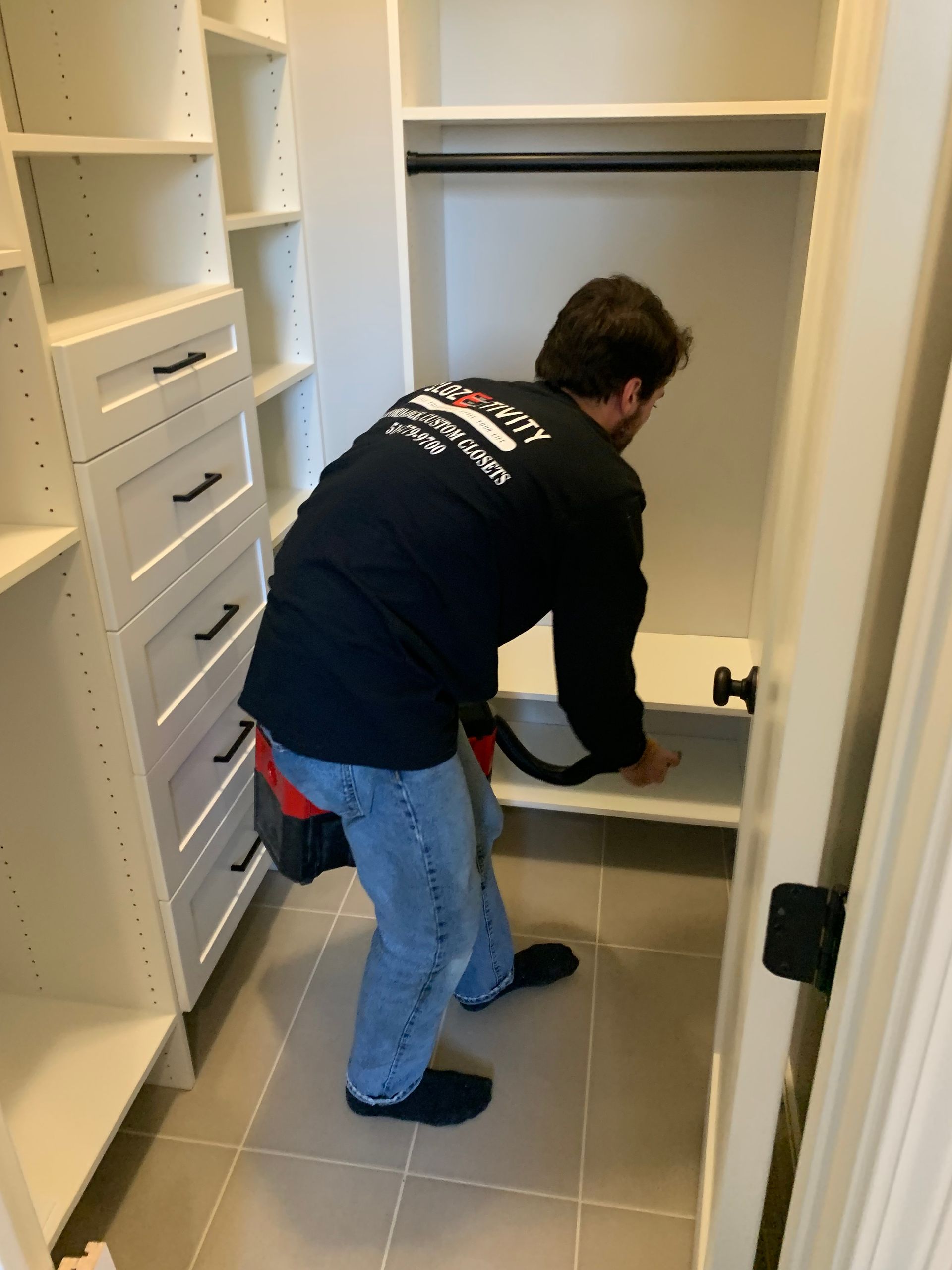 Man cleaning inside a white closet. He's wearing jeans and a black shirt, holding a vacuum.