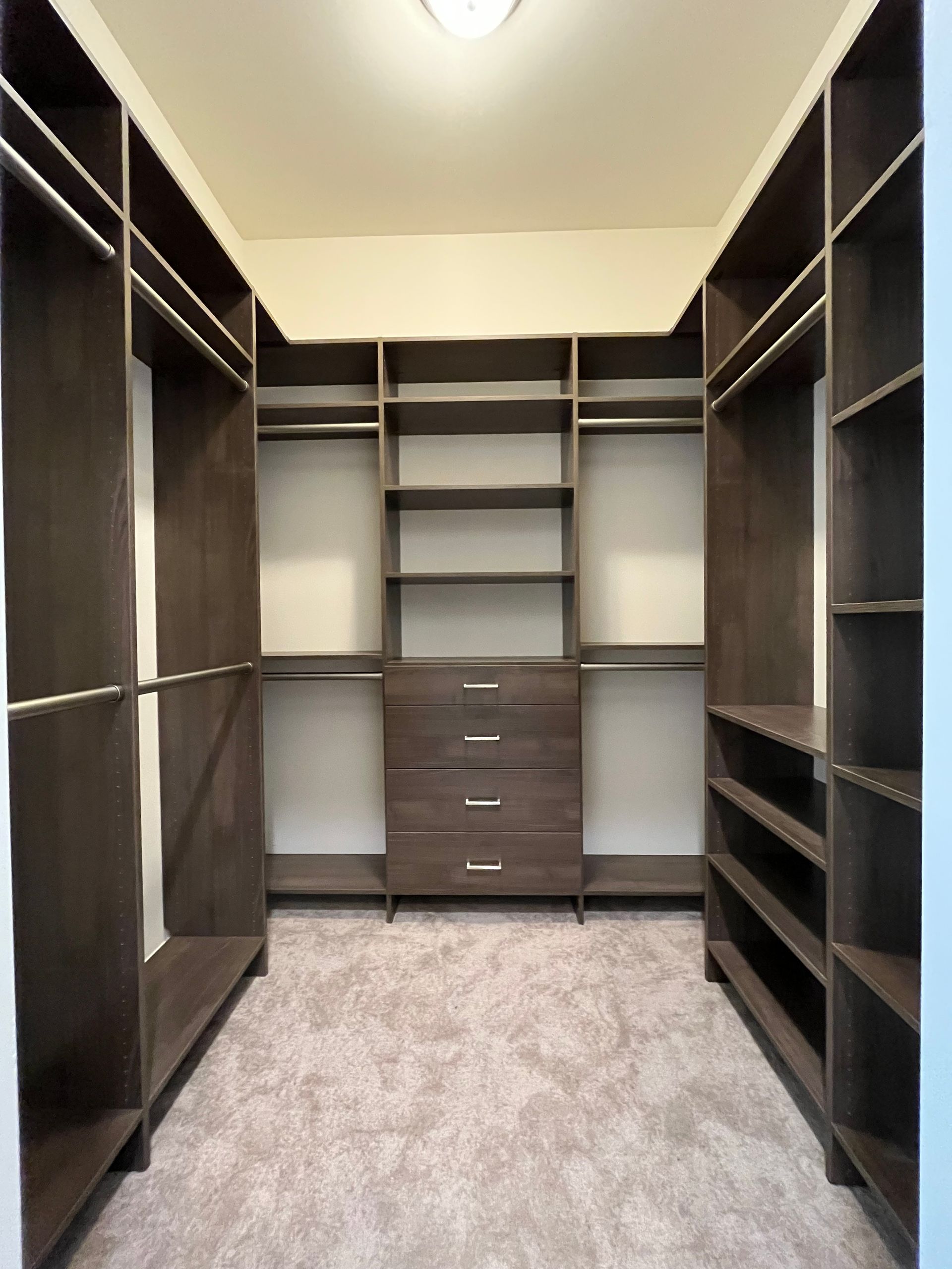 Empty dark wood walk-in closet with shelves, drawers, and hanging rods; beige walls and carpet.