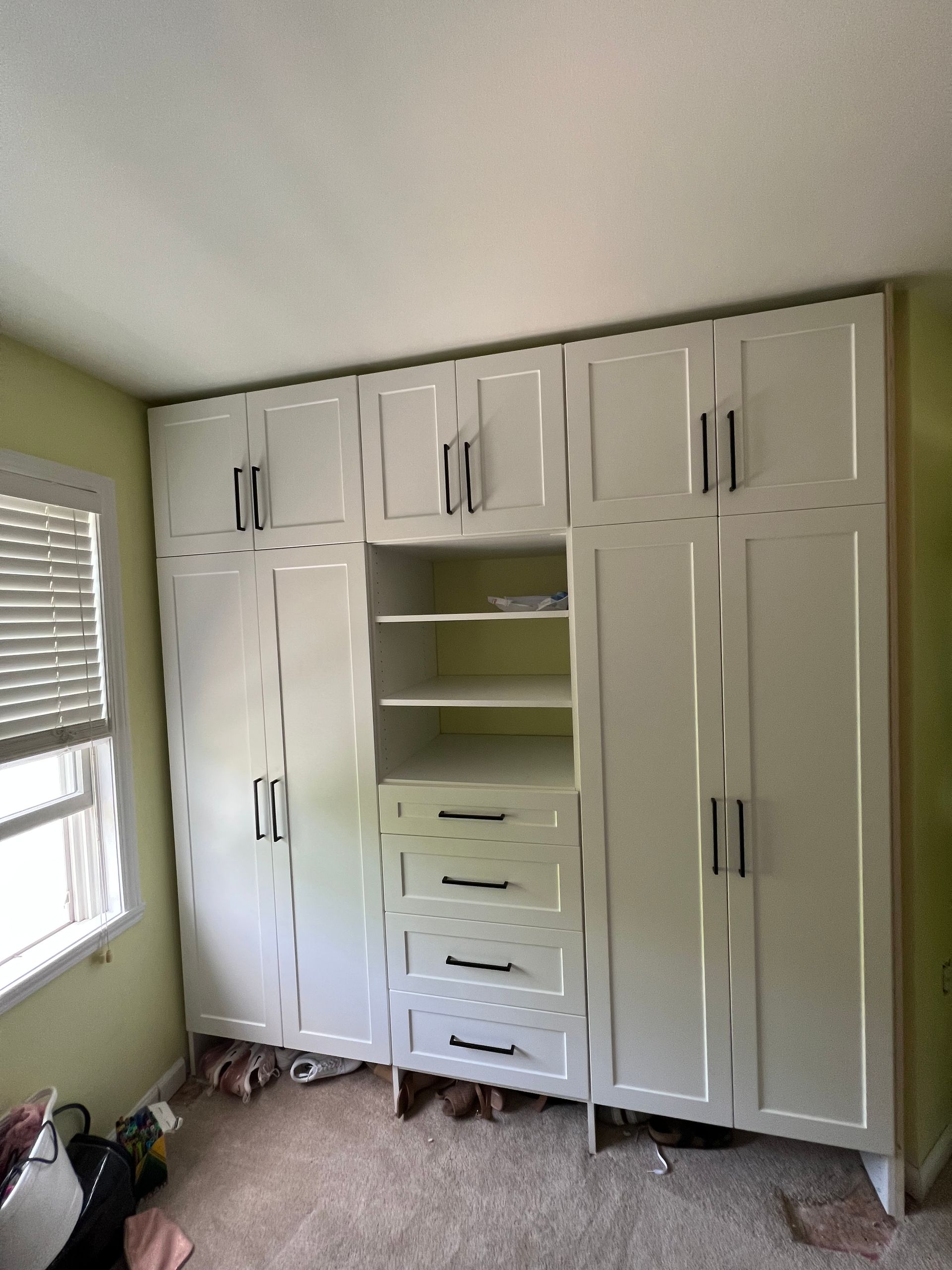 White built-in closet with drawers and shelves; black handles, in a bedroom near a window.