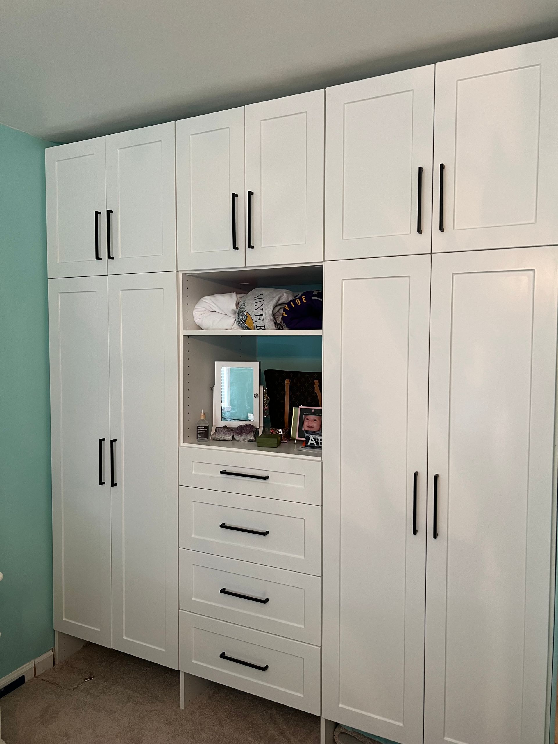 White closet with black handles against a teal wall, features cabinets, drawers, and a mirror.