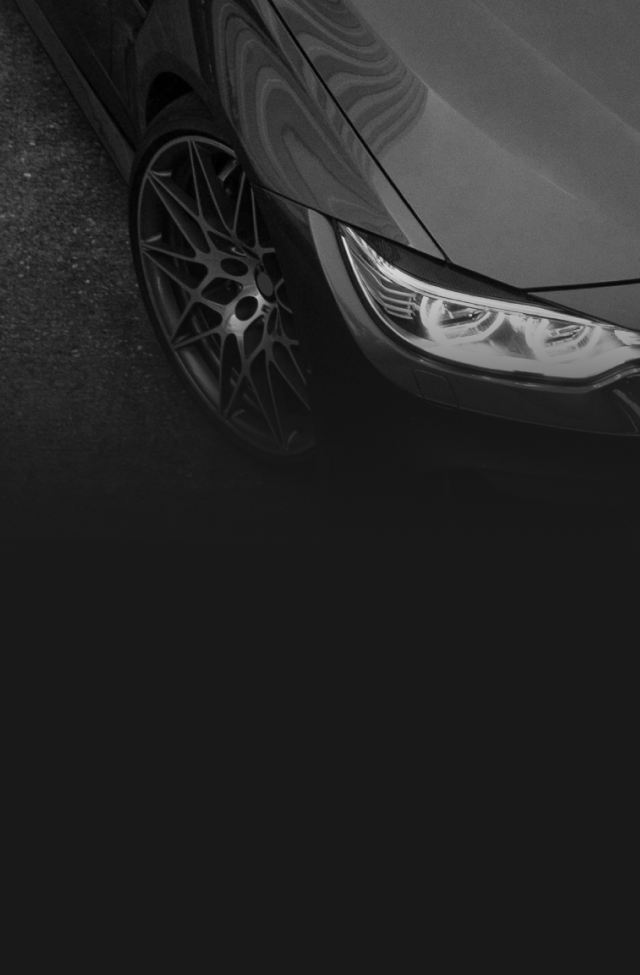 Close-up of a modern dark-colored car's front wheel and headlight, viewed from a high angle against a dark background.