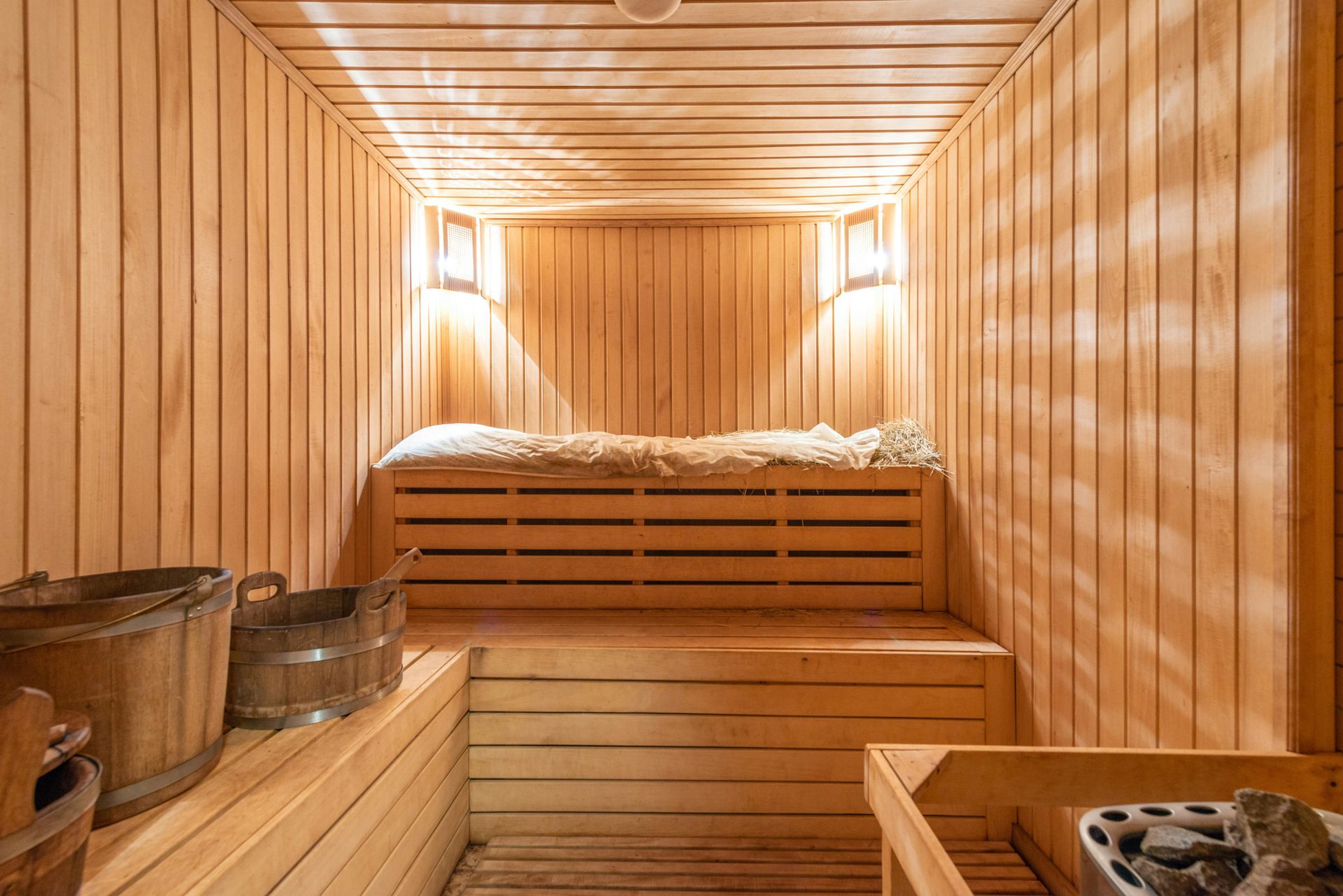 Wooden sauna interior with benches, buckets, and a heating element. Soft lighting illuminates the wood-paneled walls.